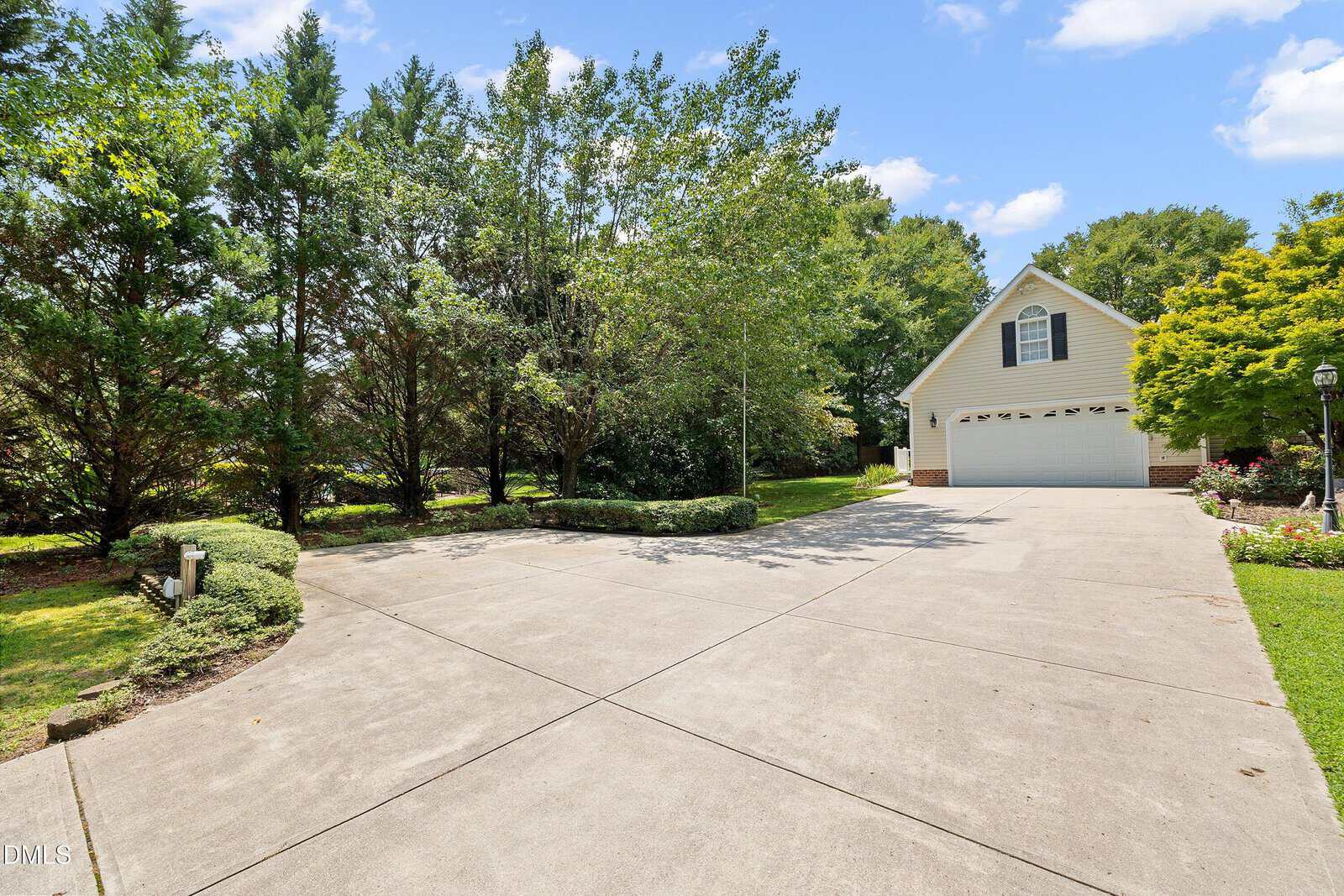 A wide concrete driveway leads to a light-colored house with a gabled roof and two windows. Trees and bushes line the driveway on both sides. The sky is mostly clear with some clouds.