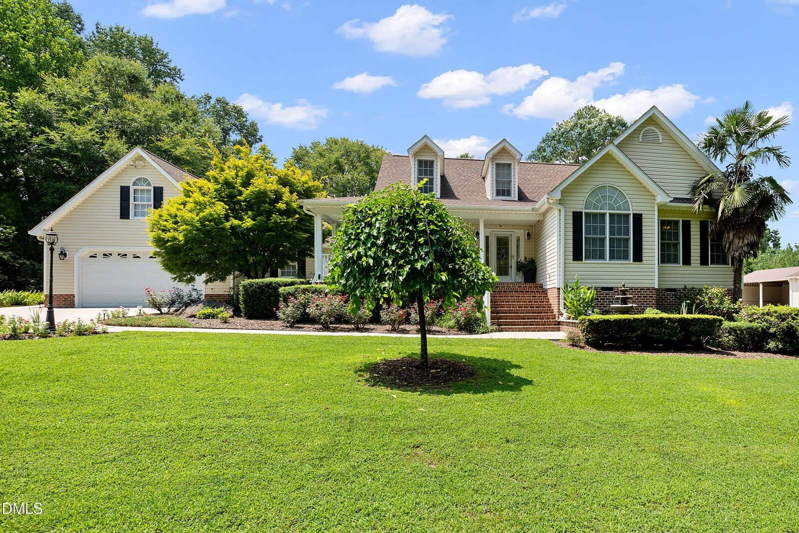 A beige suburban house with a front porch, dormer windows, and attached garage sits amid a well-manicured lawn, landscaping, and trees under a sunny blue sky with scattered clouds.
