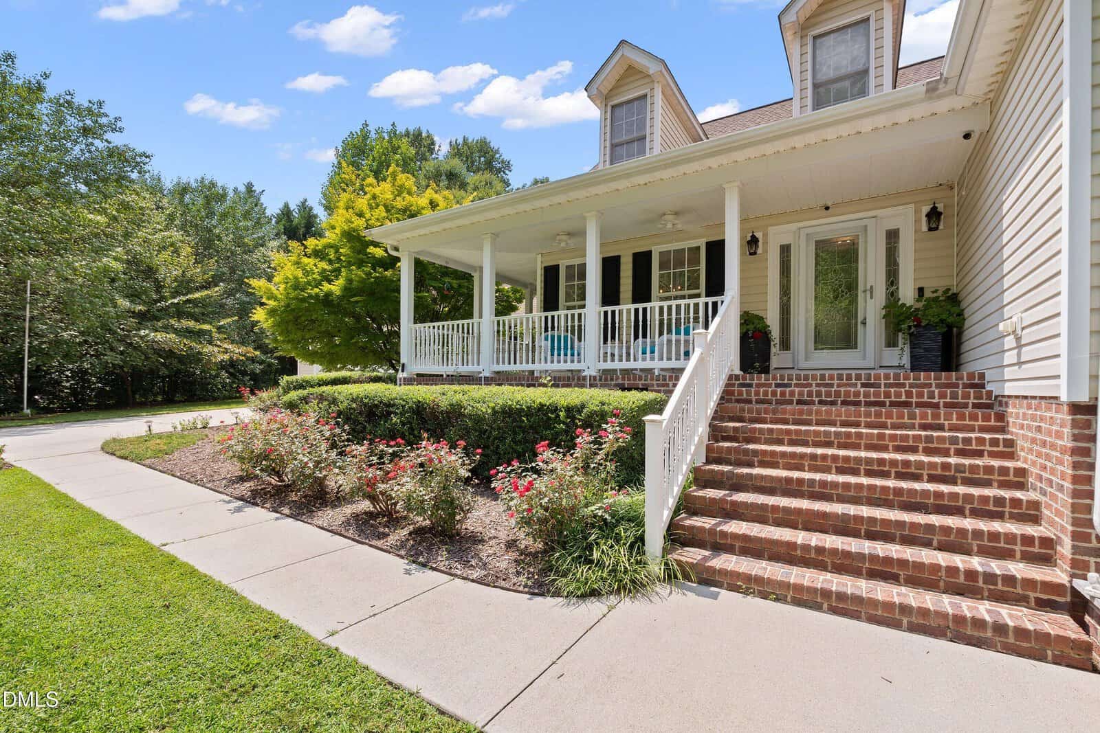 A brick stairway leads to a white porch with columns and chairs, surrounded by green lawn, shrubs, and flowers, under a partly cloudy sky.