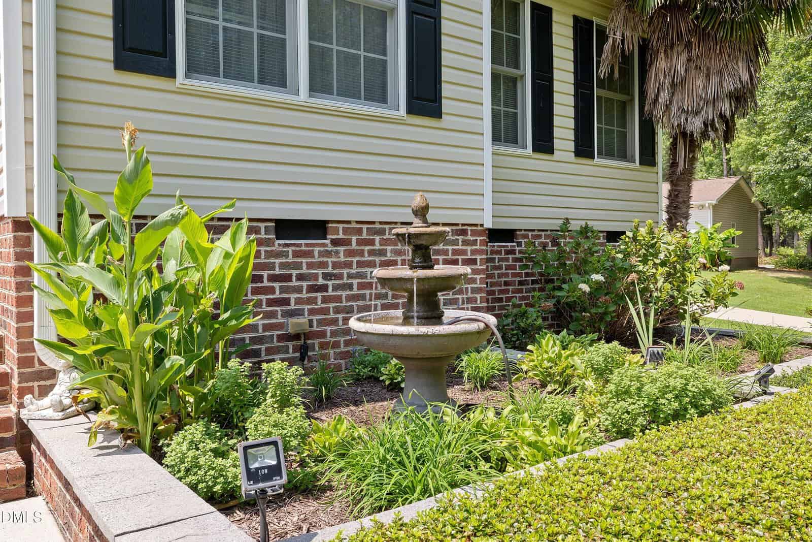 A small stone fountain stands in a landscaped garden bed with green plants, flowers, and solar lights, in front of a house with yellow siding, black shutters, and a brick foundation.