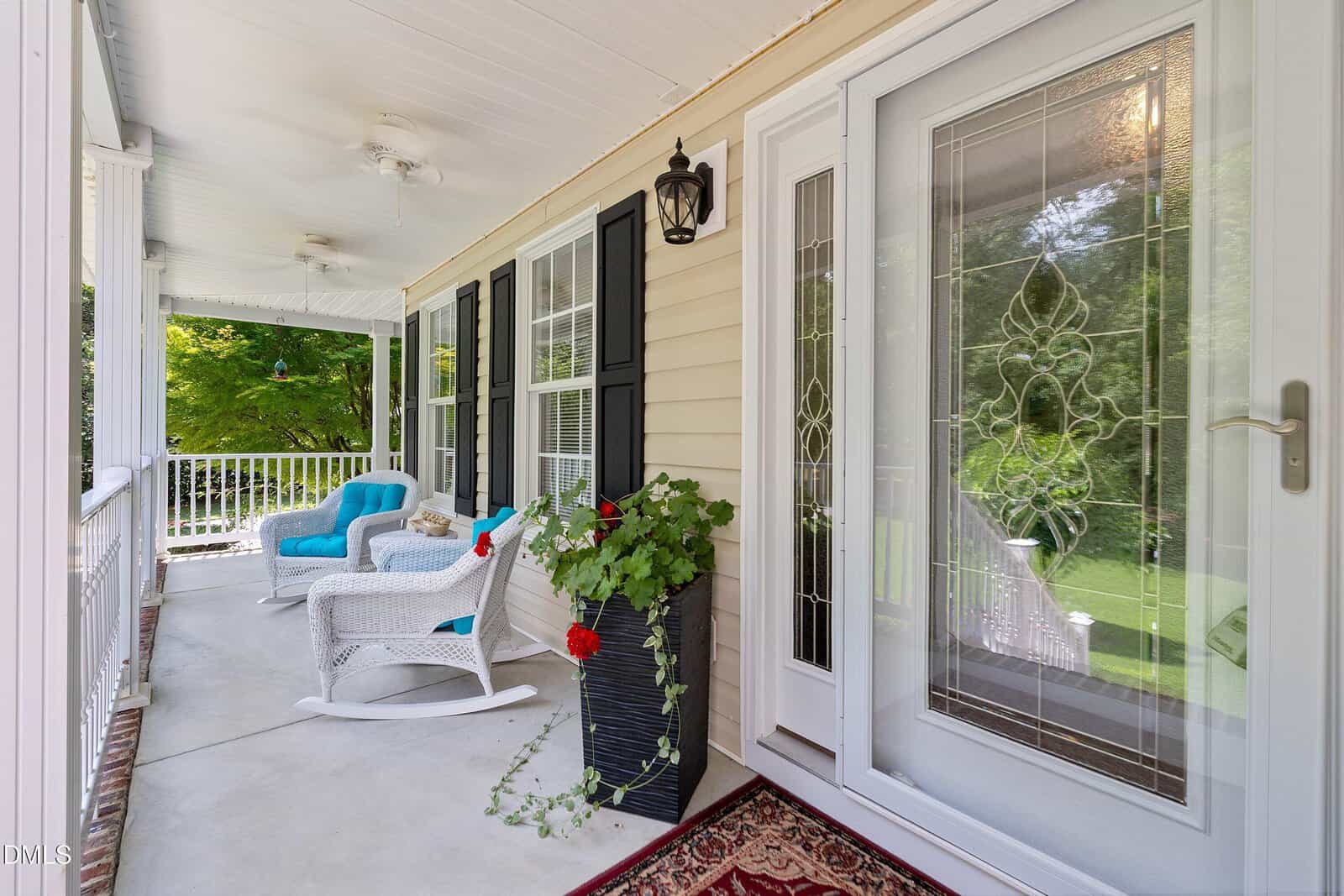 A covered porch features rocking chairs, a blue cushioned seat, potted red flowers, and a decorative glass front door. Black shutters and white railings line the house, with a green lawn visible in the background.
