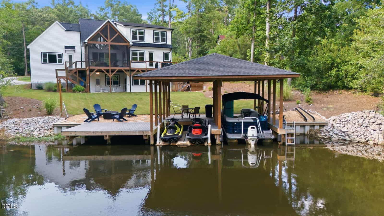 A two-story house with a covered back porch overlooks a grassy yard, fire pit area, and a dock with boats and jet skis on a calm lake, surrounded by trees and rocks.