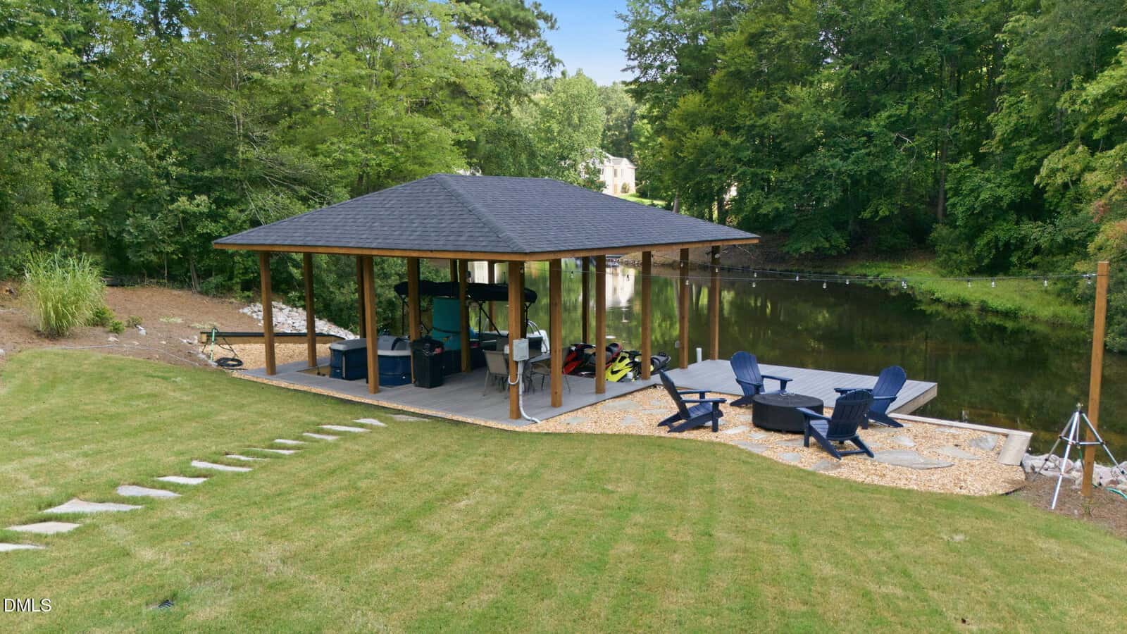 A wooden dock with a covered area sits next to a small lake. There are jet skis under the roof, Adirondack chairs around a fire pit on a deck, and a grassy lawn with stone steps leading to the water. Trees surround the scene.