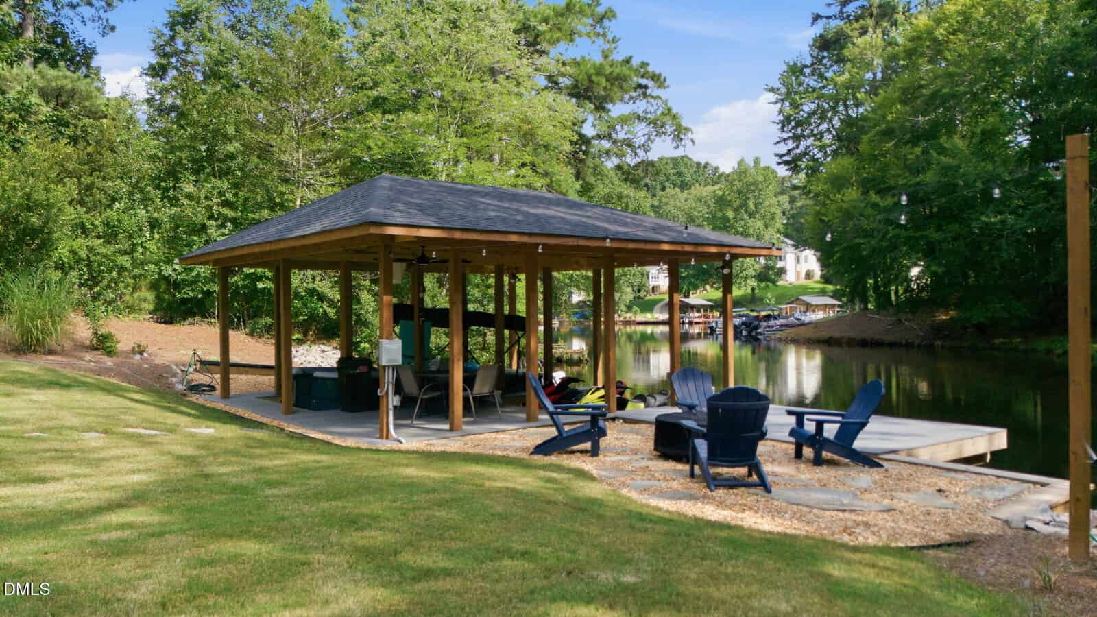 A wooden pavilion with a dark roof stands by a calm waterway, featuring outdoor seating, adirondack chairs, and a dock. Lush green trees and grass surround the area under a clear sky.