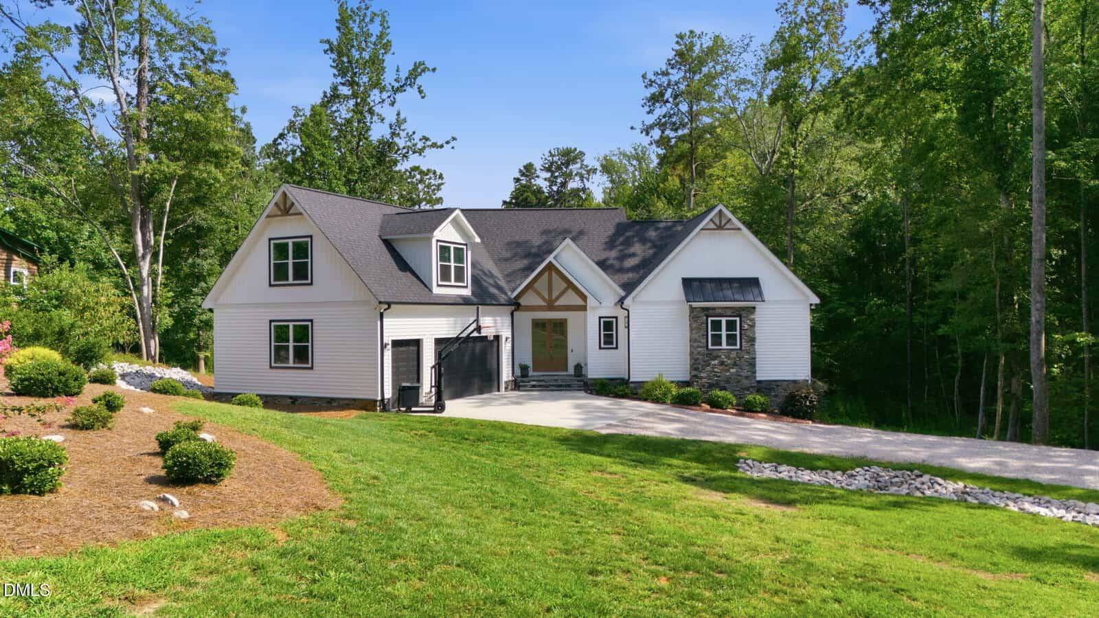 A white two-story house with dark roof shingles, stone accents, and a large driveway sits among green trees and a well-maintained lawn on a sunny day.