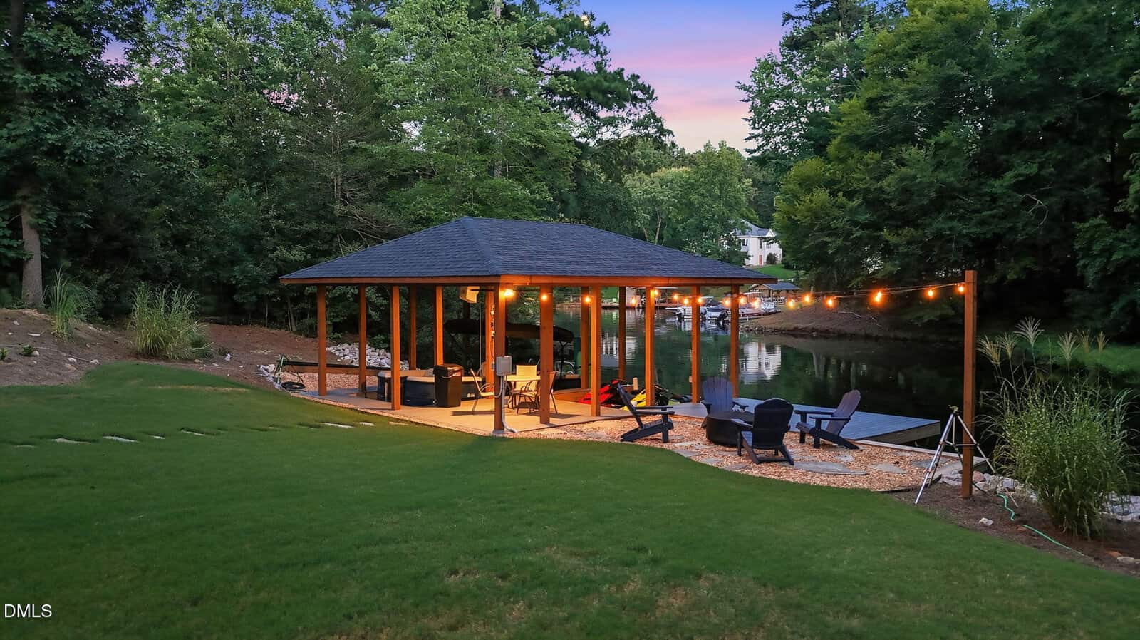 A covered outdoor patio by a lake at dusk, with string lights, chairs, and a fire pit, surrounded by green grass and tall trees.