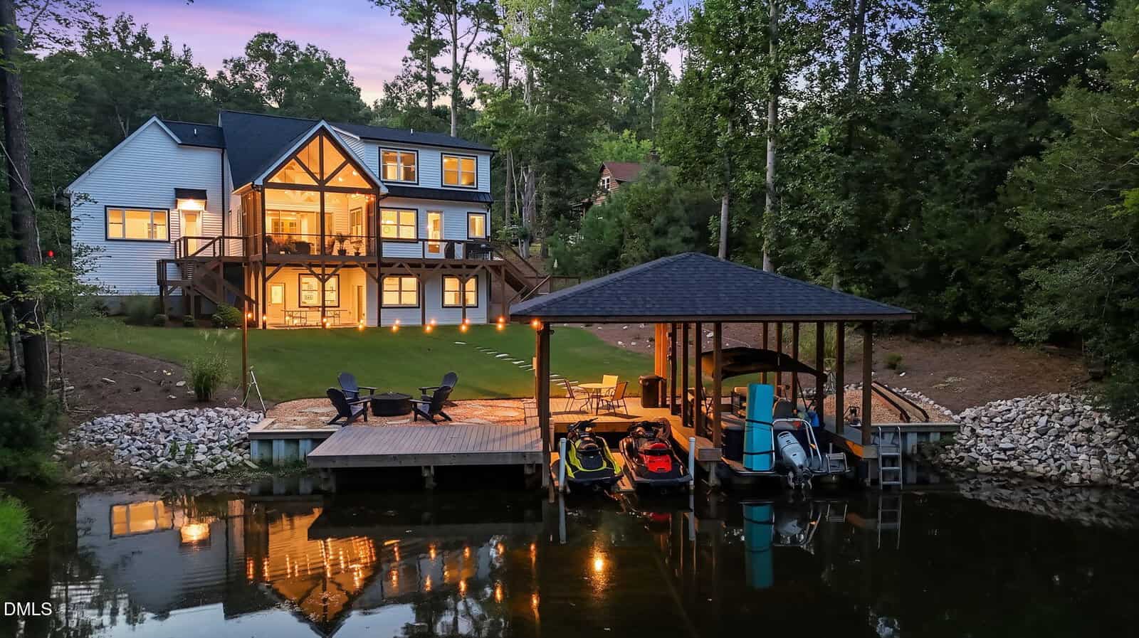 A modern lakeside house at 683 Sagamore Drive, Louisburg, with large windows and decks, is lit up at sunset. A covered dock with boats and jet skis sits on calm water, surrounded by trees and a grassy yard.