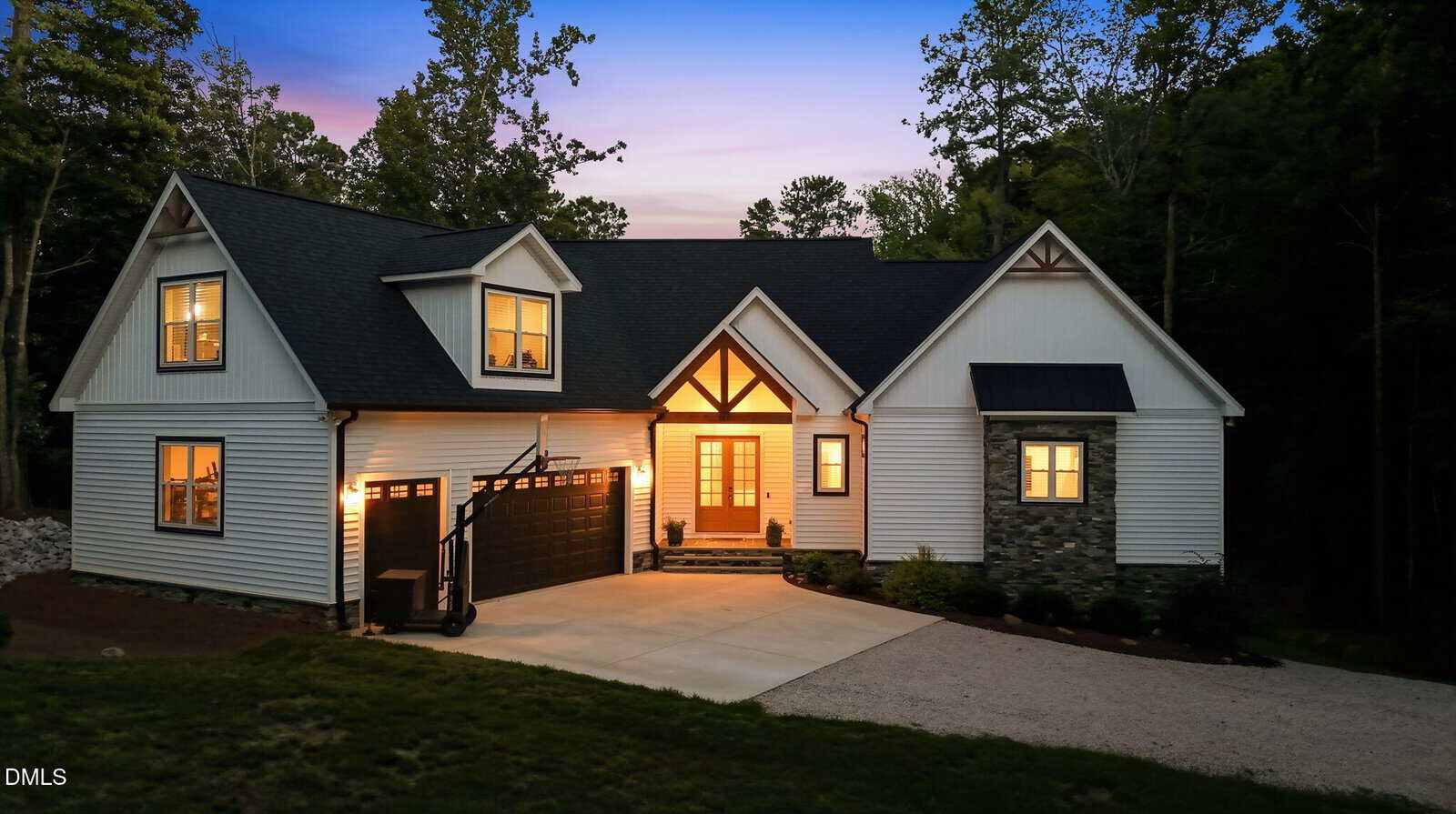 A modern two-story house with lit windows at dusk, black garage door, white siding, stone accents, and a driveway, surrounded by trees.