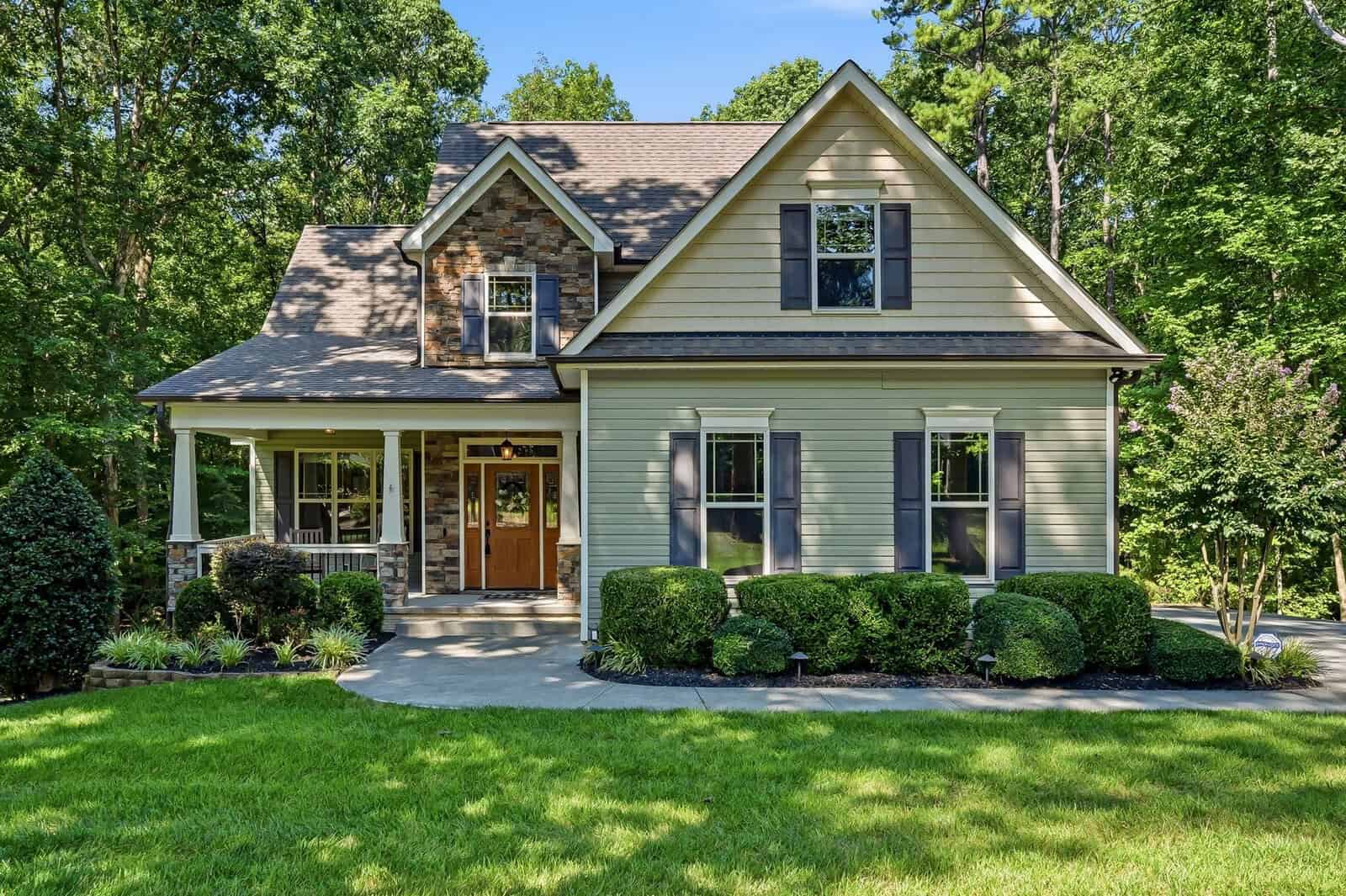 A two-story beige house with black shutters, a wooden front door, and a covered porch. It is surrounded by green shrubs, a manicured lawn, and tall trees in the background under a clear blue sky.