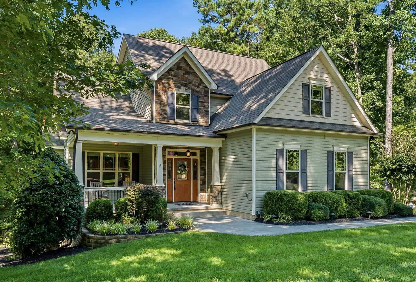 Two-story house with beige siding, gray shutters, and stone accents. Front porch with columns, landscaped yard, shrubs, and trees. Concrete walkway leads to the wooden front door. Lush grass and forested background.
