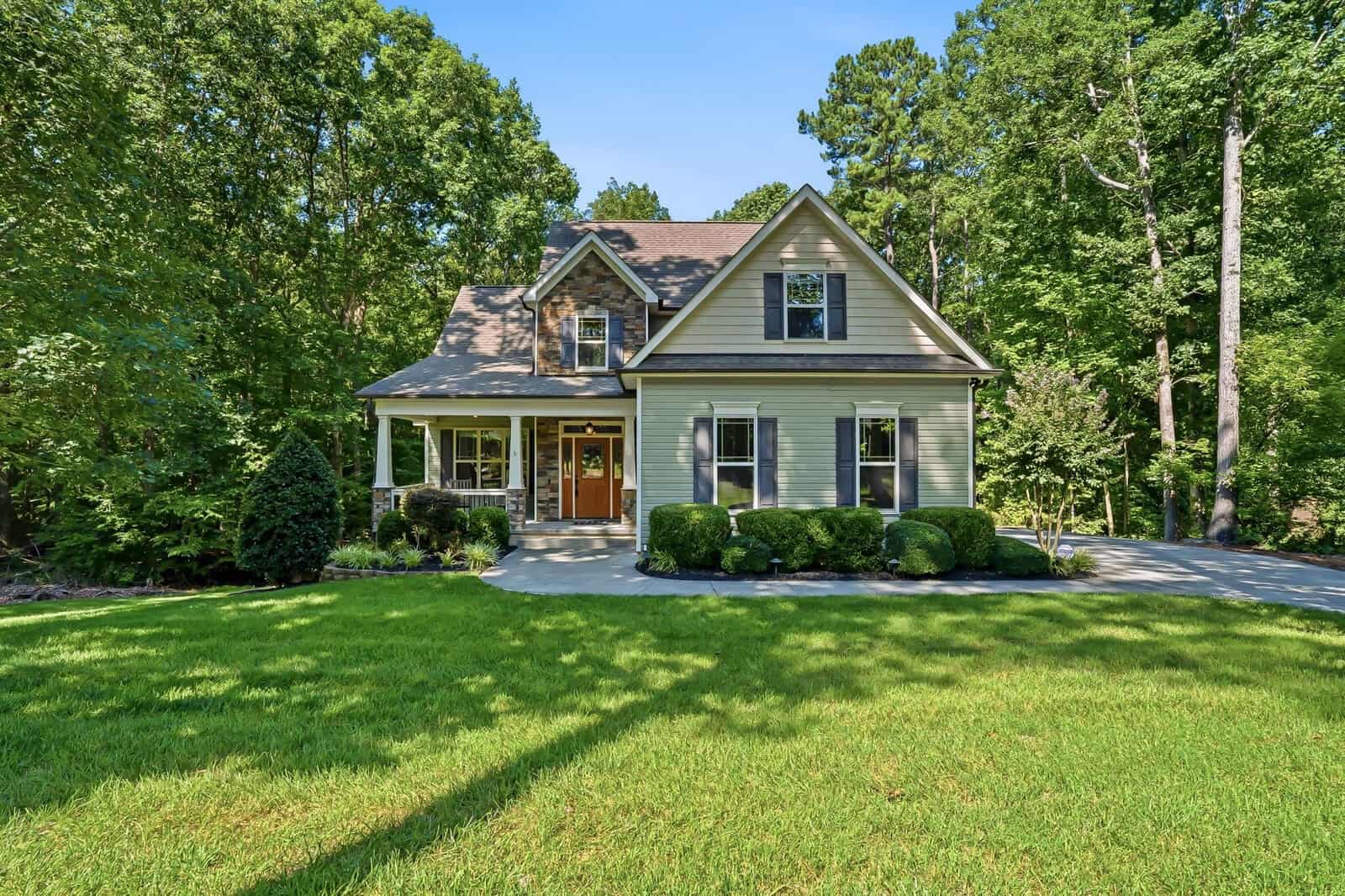 Two-story suburban house with gray siding, white trim, and a covered front porch. The home is surrounded by lush green grass, bushes, and tall trees under a clear blue sky.
