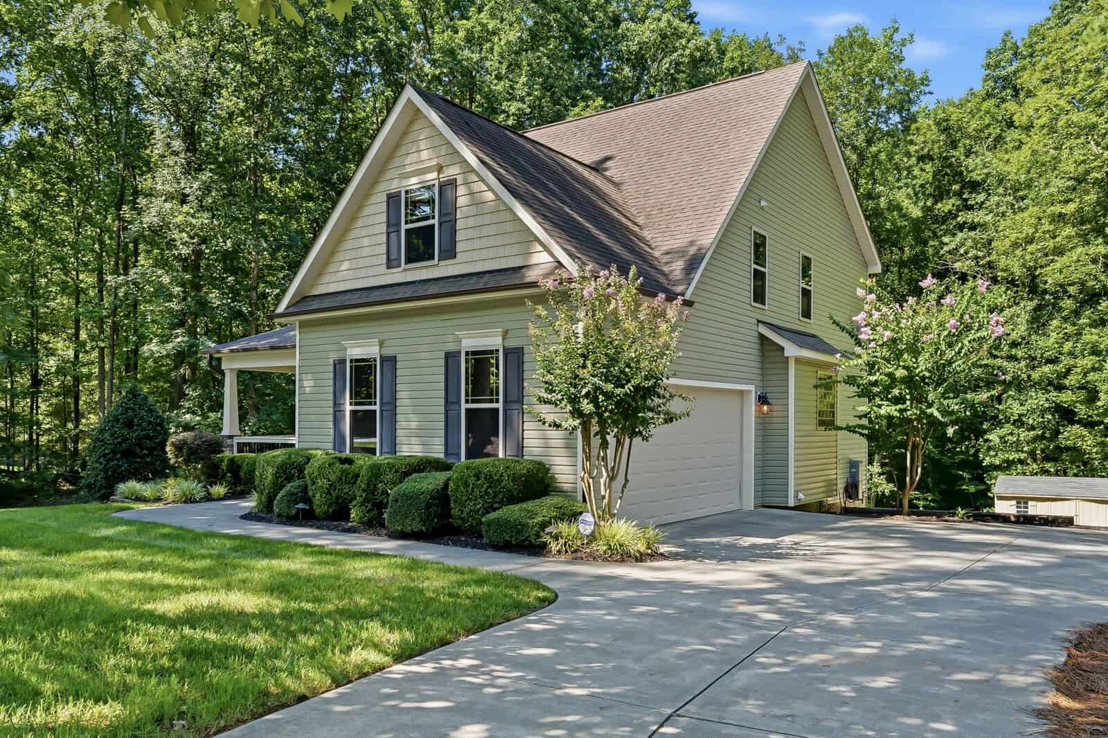 Two-story beige house with a gabled roof and attached two-car garage. The house is surrounded by trimmed bushes, green grass, and trees in the background, with a concrete driveway in the foreground.