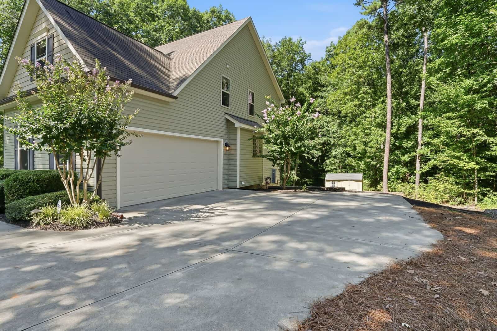 A beige house with a two-car garage, sloped roof, and large driveway surrounded by trees and landscaping on a sunny day.
