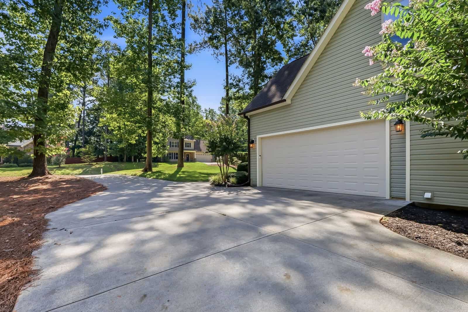 A wide concrete driveway leads to a two-car garage attached to a light green house, surrounded by tall trees and green lawn on a sunny day.