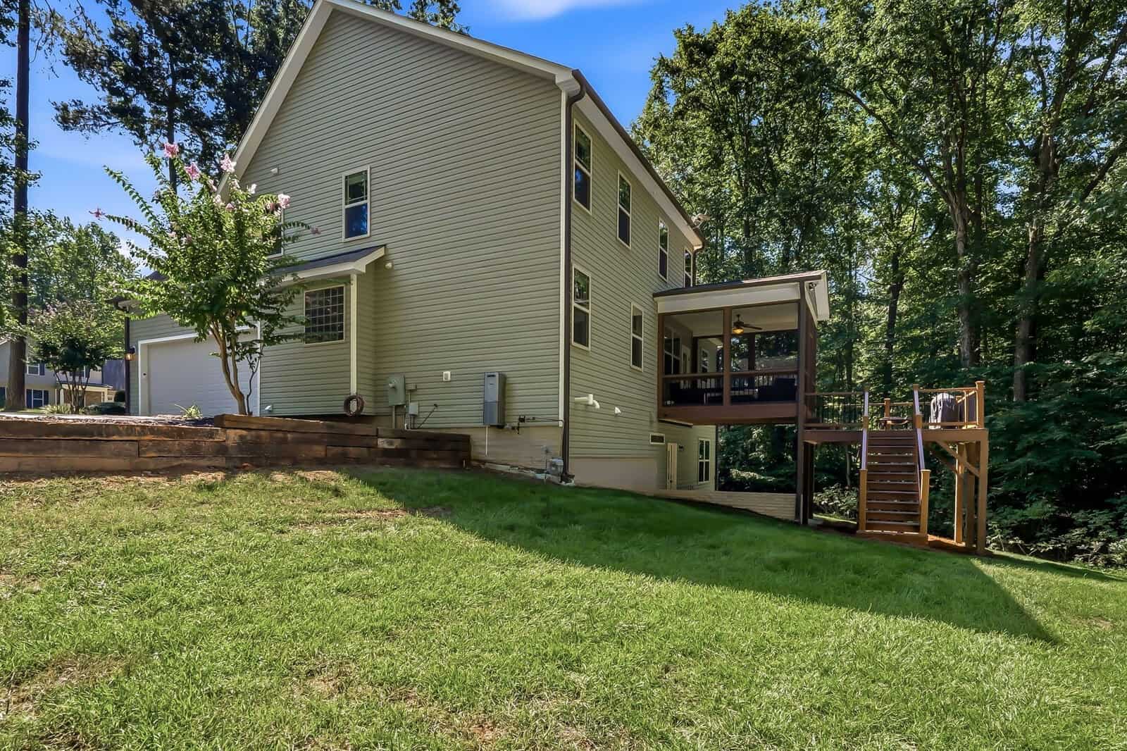 Two-story beige house with a screened porch, wooden deck, and steps leading to a grassy backyard. Surrounded by trees and a wooden retaining wall on the left. The sky is clear and blue.