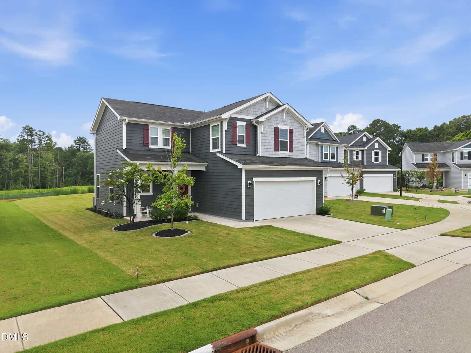 Two-story house with gray siding, white trim, and red shutters, featuring a double garage and a manicured lawn. The home is located in a suburban neighborhood with similar houses under a partly cloudy sky.