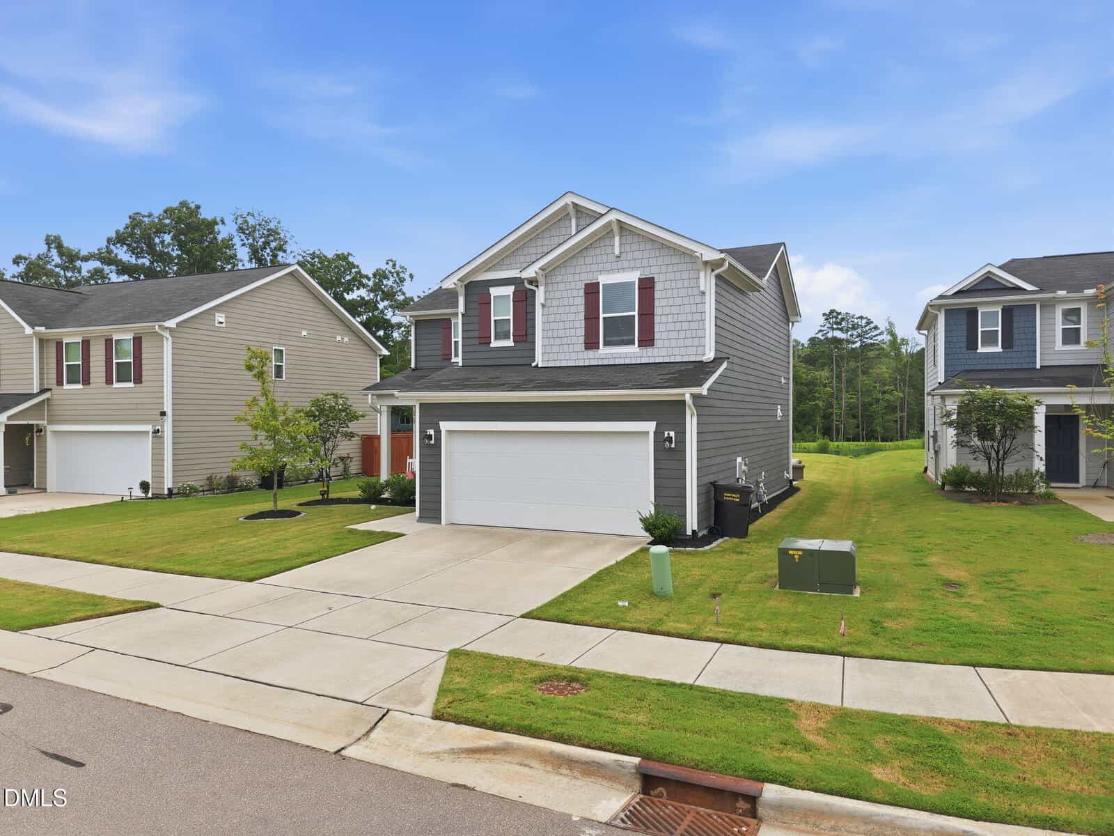 Two-story gray house with white trim, a double garage, red shutters, and a well-kept green lawn. The house is in a suburban neighborhood with similar homes and trees in the background under a blue sky.