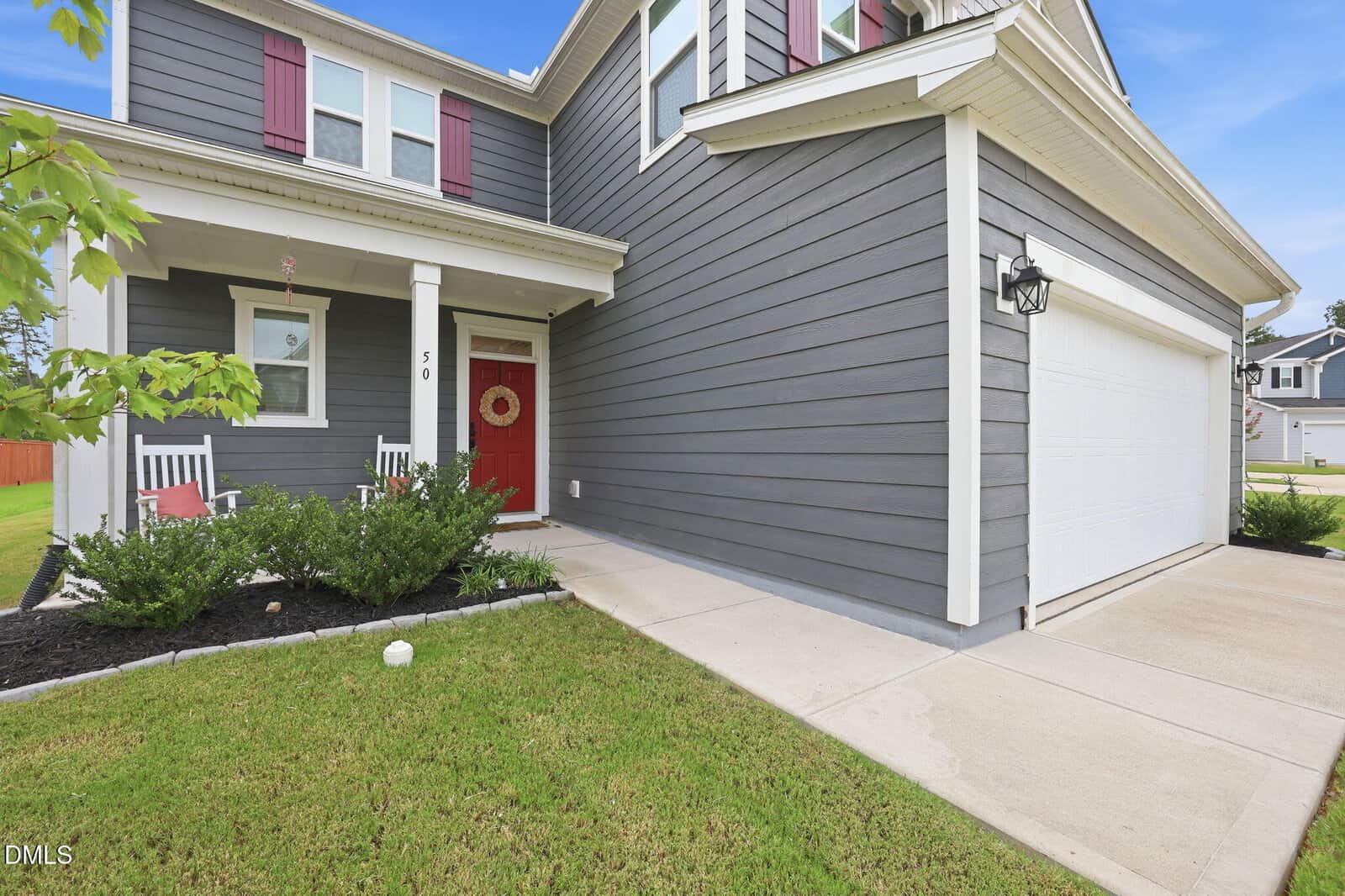 Two-story gray house with white trim, red front door, white garage door, covered porch with two white rocking chairs, landscaping with green bushes, and a lawn bordered by a concrete driveway and sidewalk.