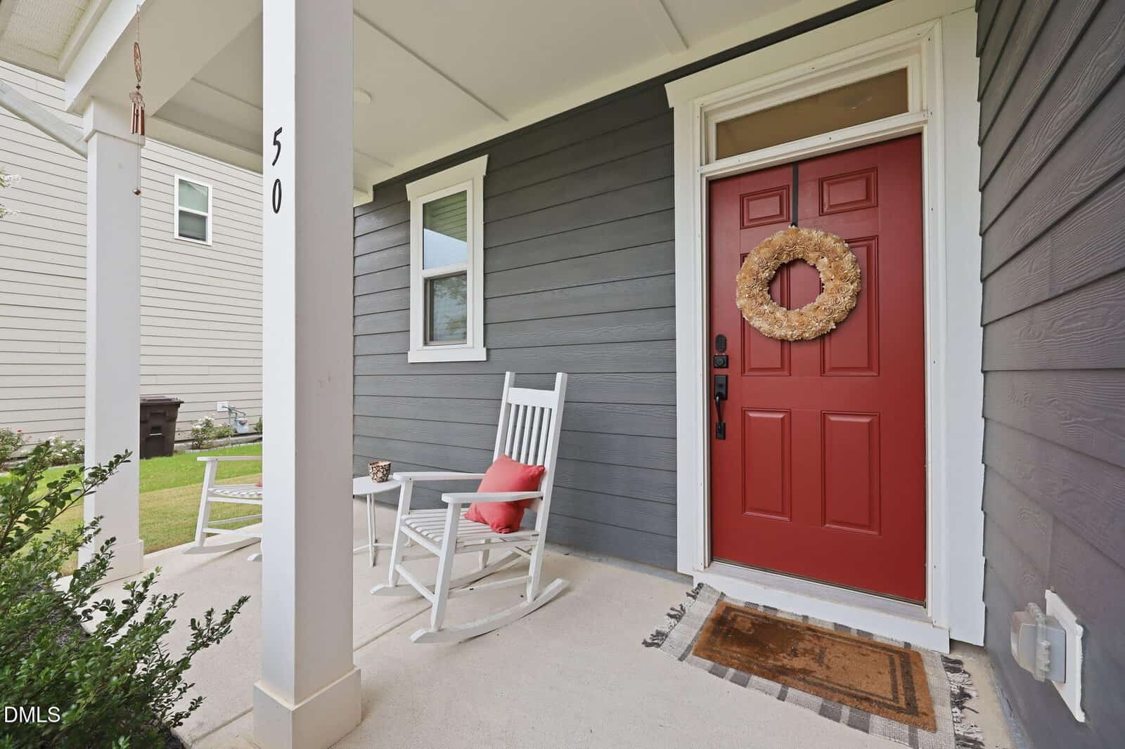 Gray house exterior with a red front door, a wreath hanging on the door, a doormat, and two white rocking chairs with red pillows on a covered porch. A small side table is between the chairs. House number 50 is visible.