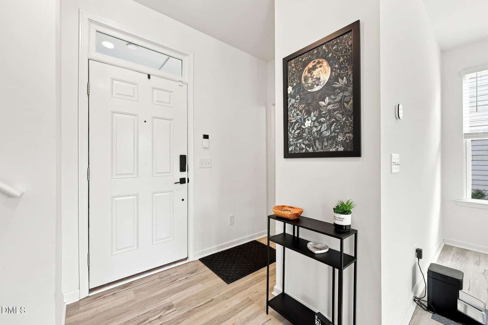 Bright entryway with white walls, a white front door, light wood floors, a black console table holding small decor, and a framed botanical moon artwork hanging above the table.