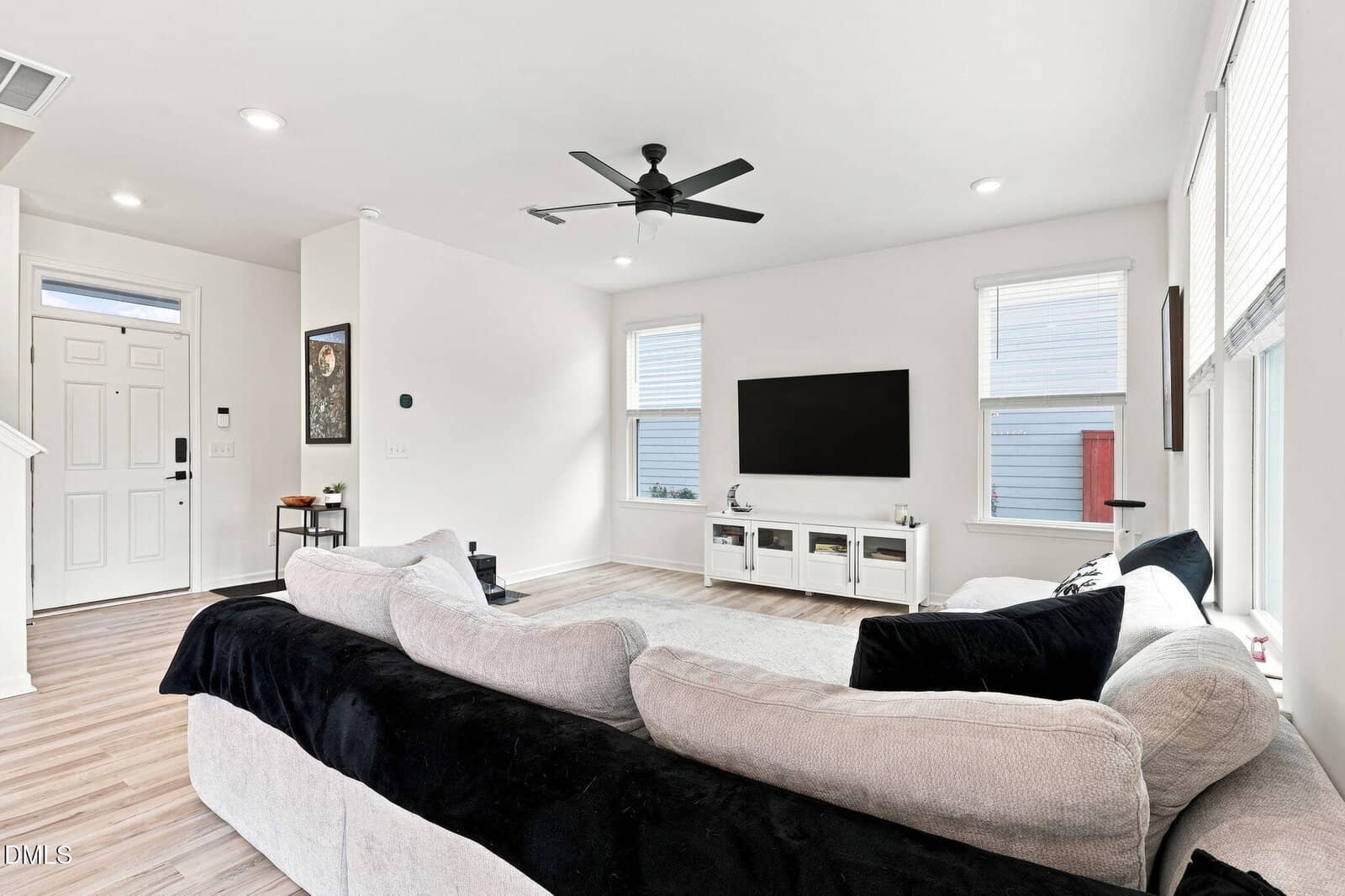 Bright living room with a large sectional sofa, wall-mounted TV, white TV stand, light wood flooring, and several windows letting in natural light. A ceiling fan is centered on the white ceiling.