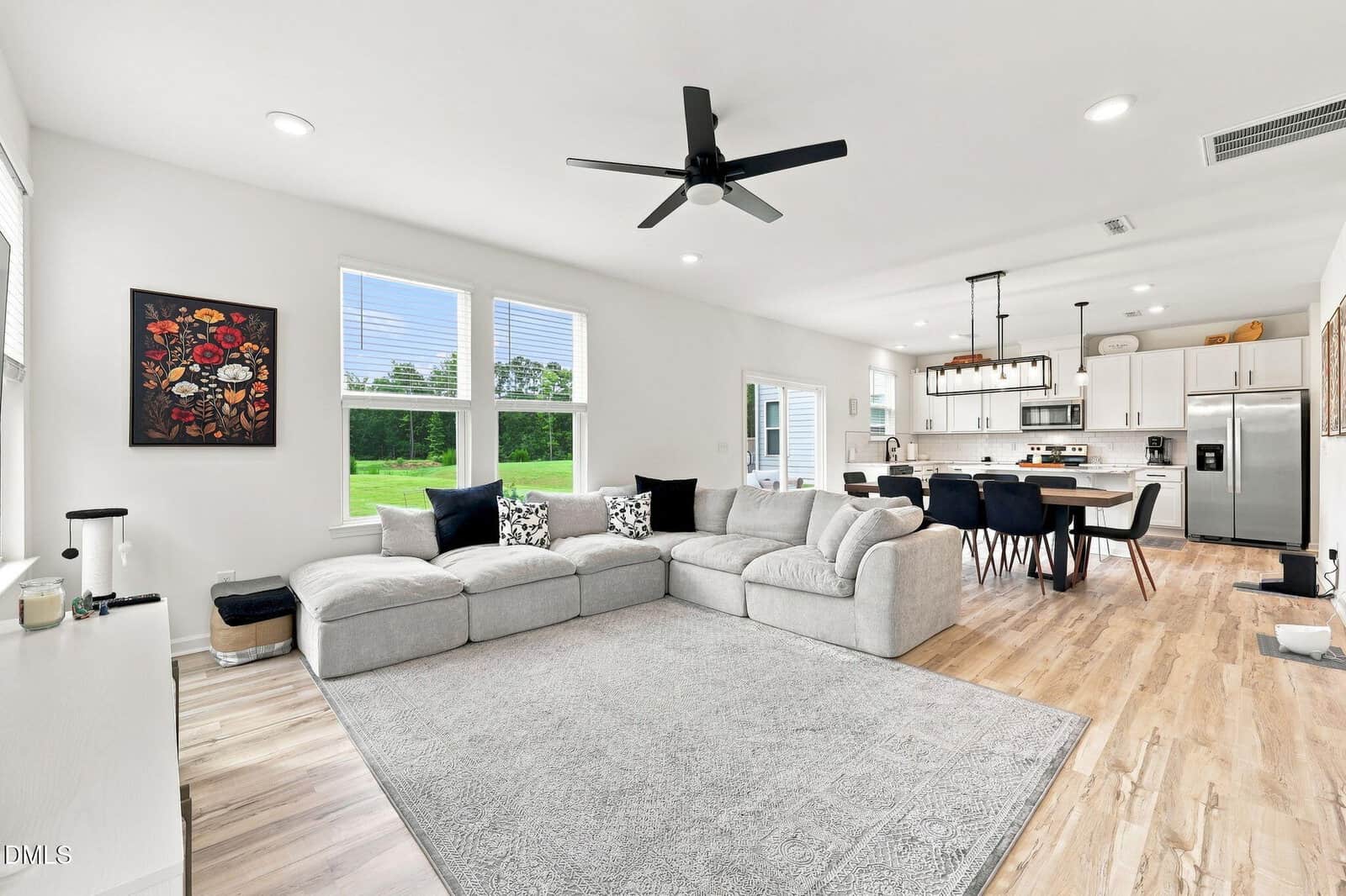 Modern open-concept living room and kitchen with light wood floors, a gray sectional sofa, dining table with black chairs, stainless steel appliances, ceiling fan, large windows, and wall art.
