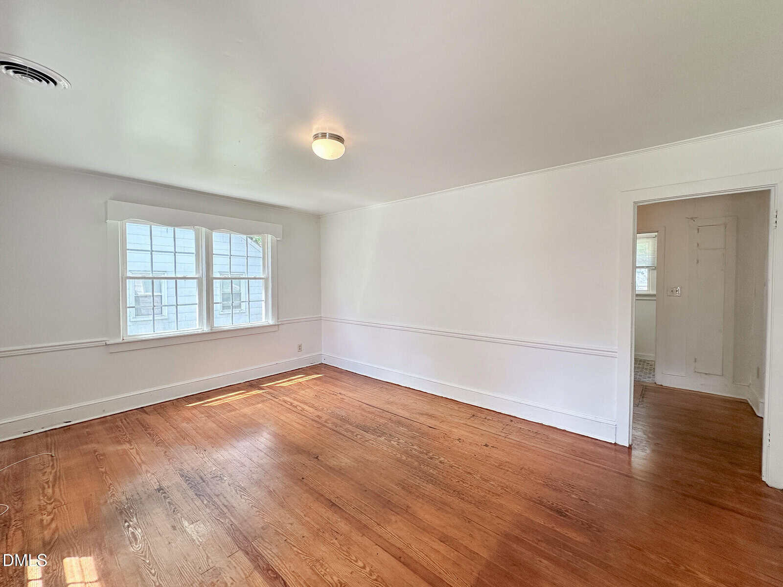 Bright, empty room with white walls, a ceiling light, large window letting in natural light, hardwood floors, and a doorway leading to another area of the home.