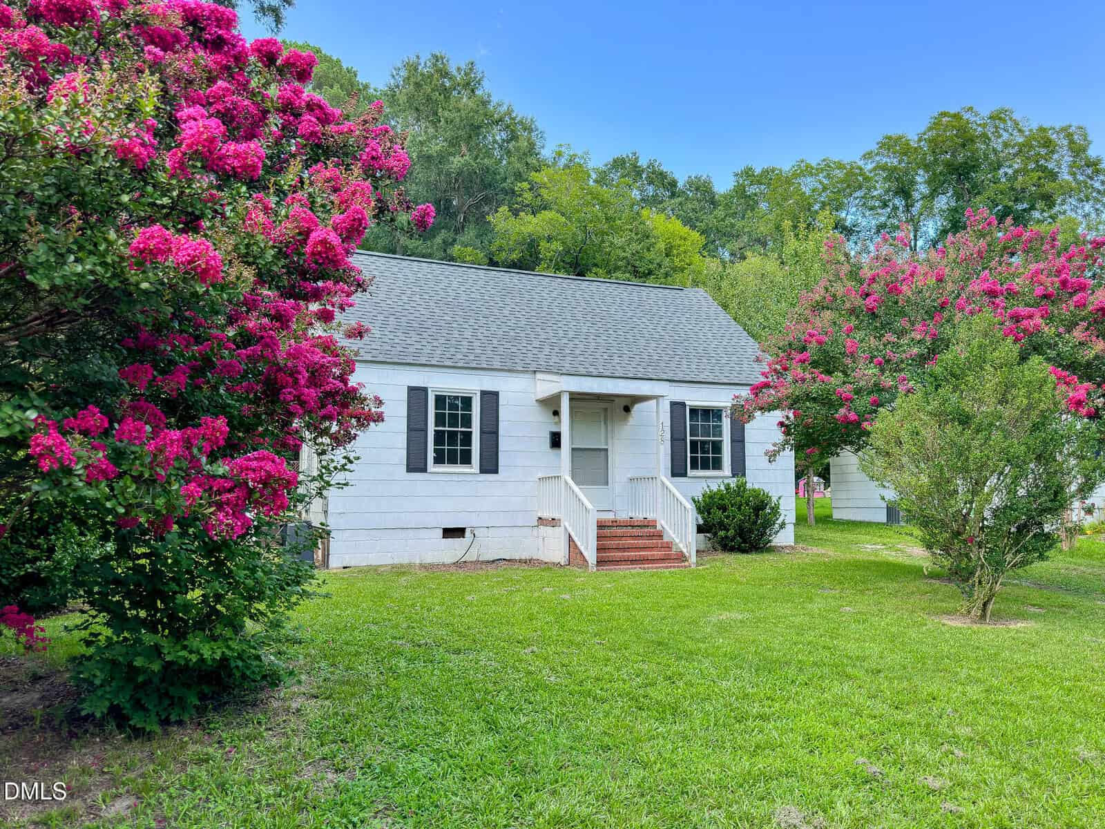 Charming small white house with gray roof at 128 Battle Avenue, Warrenton, featuring front steps and a welcoming porch surrounded by a green lawn and blooming pink crepe myrtle trees under a clear blue sky.