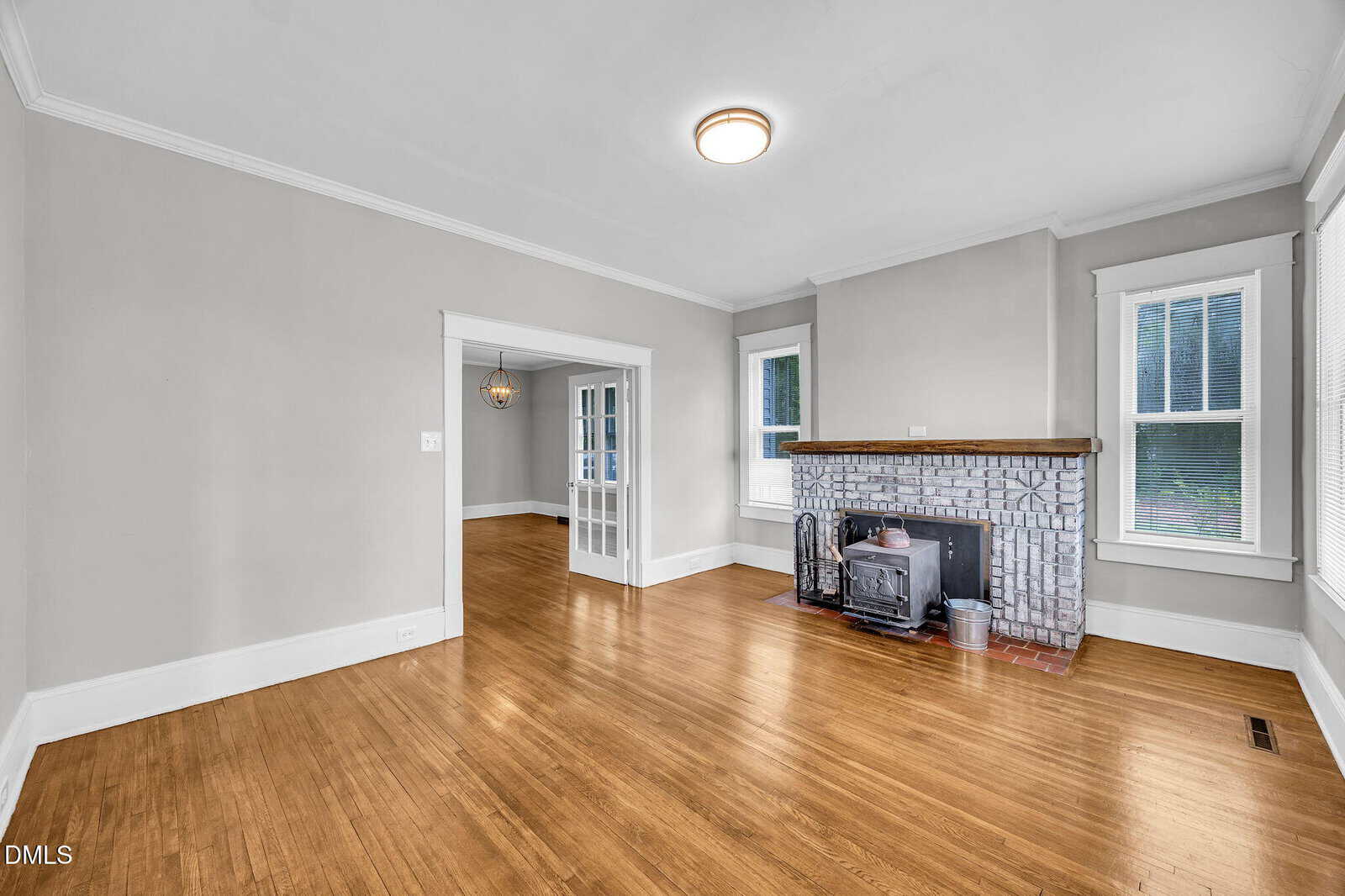 Unfurnished living room with light gray walls, wood floor, brick fireplace with wood stove, white trim, two windows, and a ceiling light. An open doorway leads to an adjoining room with glass-paneled doors.