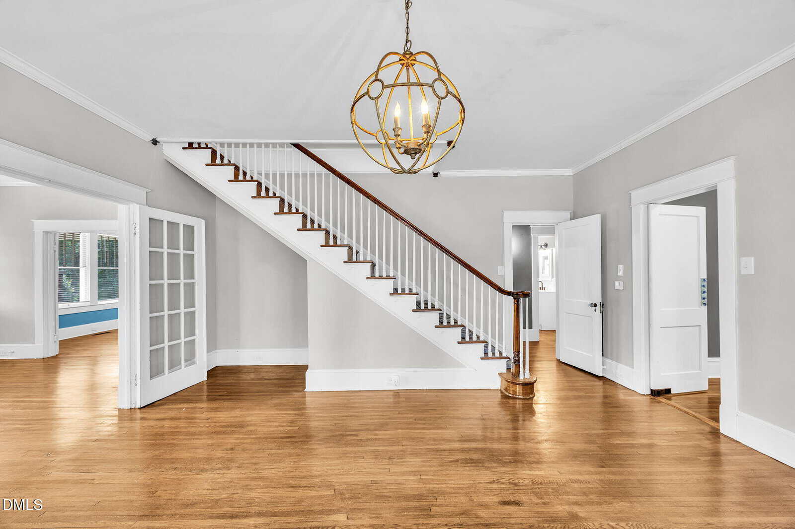 A room with hardwood floors, white walls, a wooden staircase with white spindles, a gold chandelier, and multiple doorways leading to adjacent rooms with visible natural light.