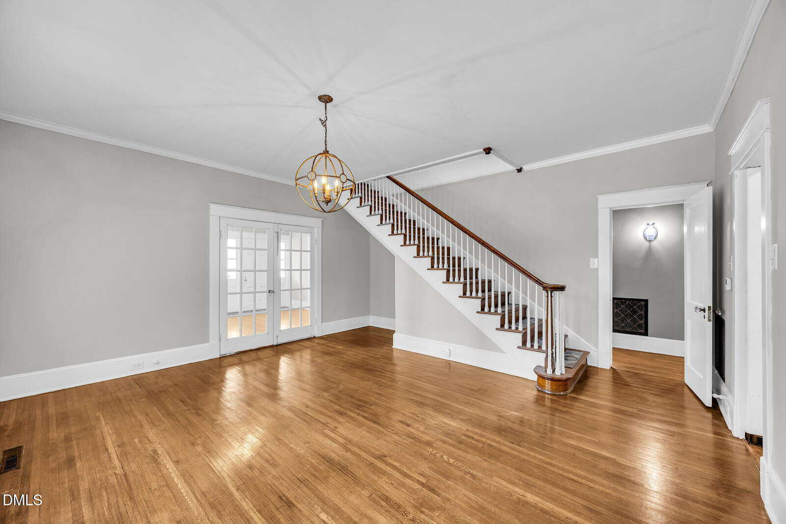 Empty living room with light gray walls, hardwood floors, a staircase with wooden railing, a round ceiling light fixture, and double glass doors leading to another room. White trim outlines windows, doors, and baseboards.