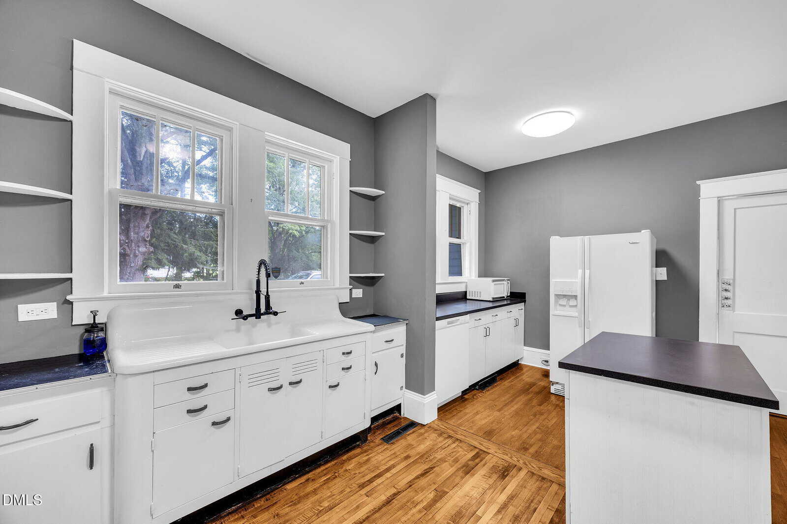 Kitchen with white cabinets, a vintage white sink, open shelves, black countertops, a white refrigerator, microwave, wood floors, and gray walls. Two windows provide natural light. Ceiling light and door visible in the background.