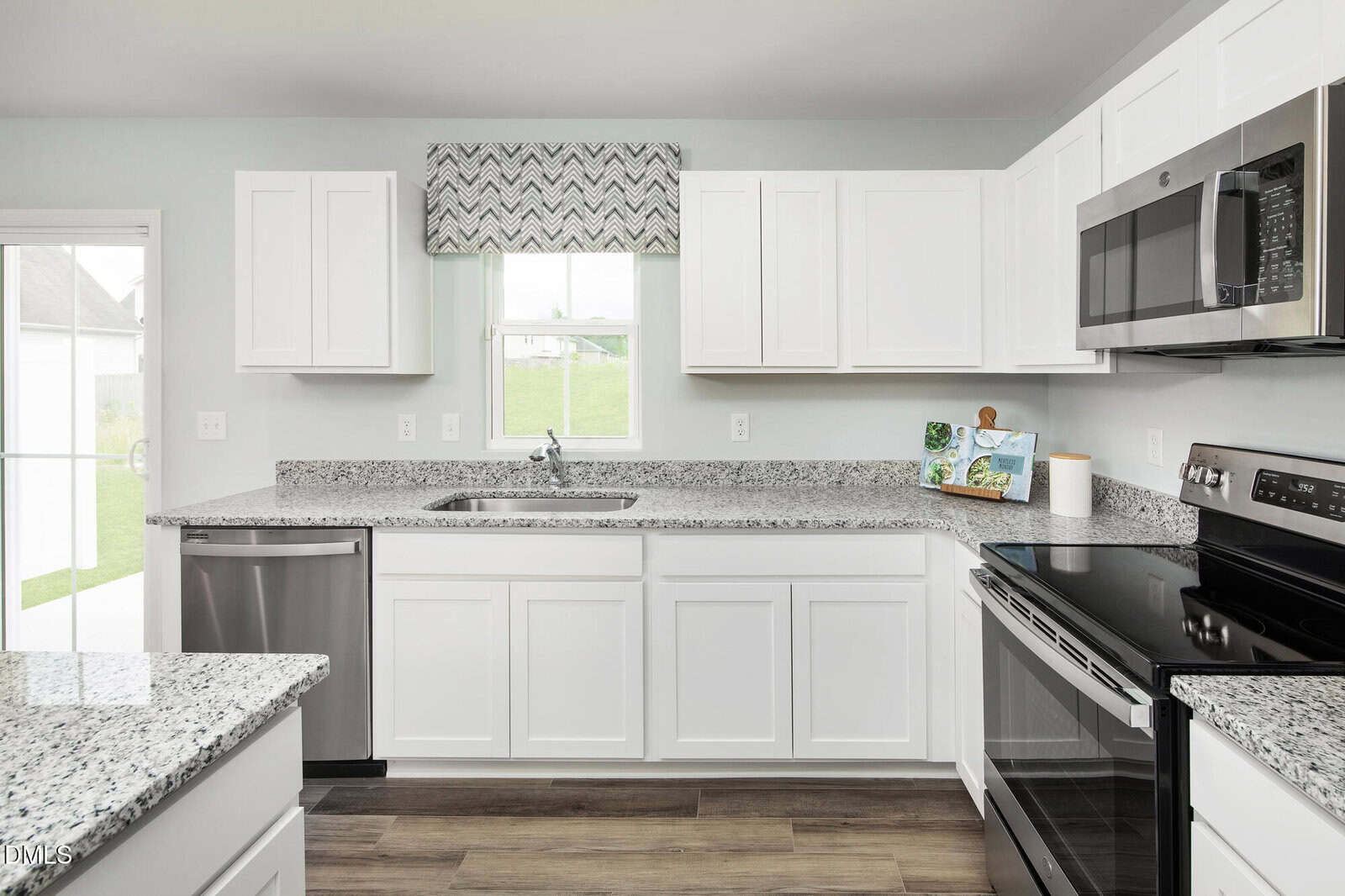 Modern kitchen with white cabinets, granite countertops, stainless steel appliances, a window with a patterned valance above the sink, and a view of a lawn outside. Wooden flooring and neutral walls complete the space.