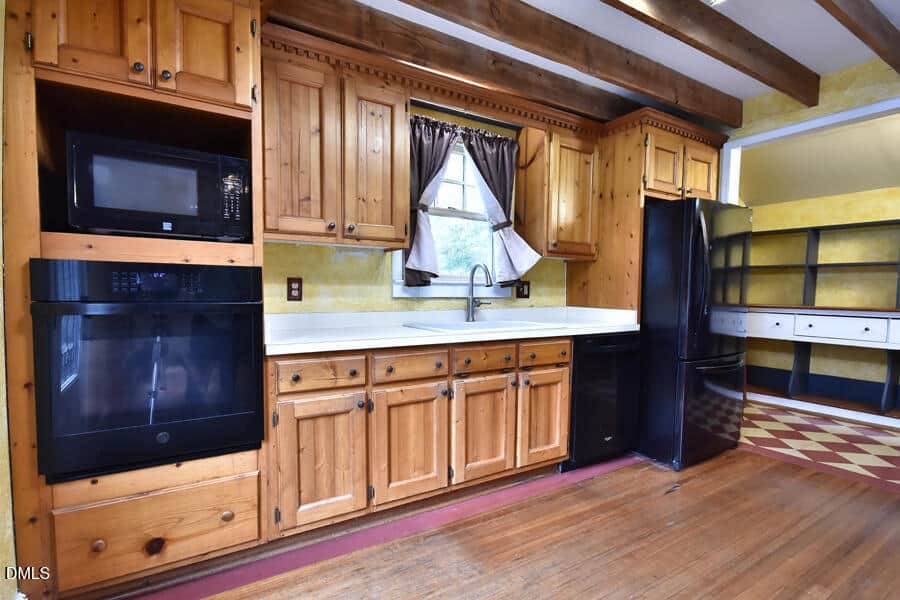 Rustic kitchen with wooden cabinets, white countertops, a black fridge, oven, microwave, and a sink under a window with brown curtains. Yellow walls and wooden beams decorate the space.
