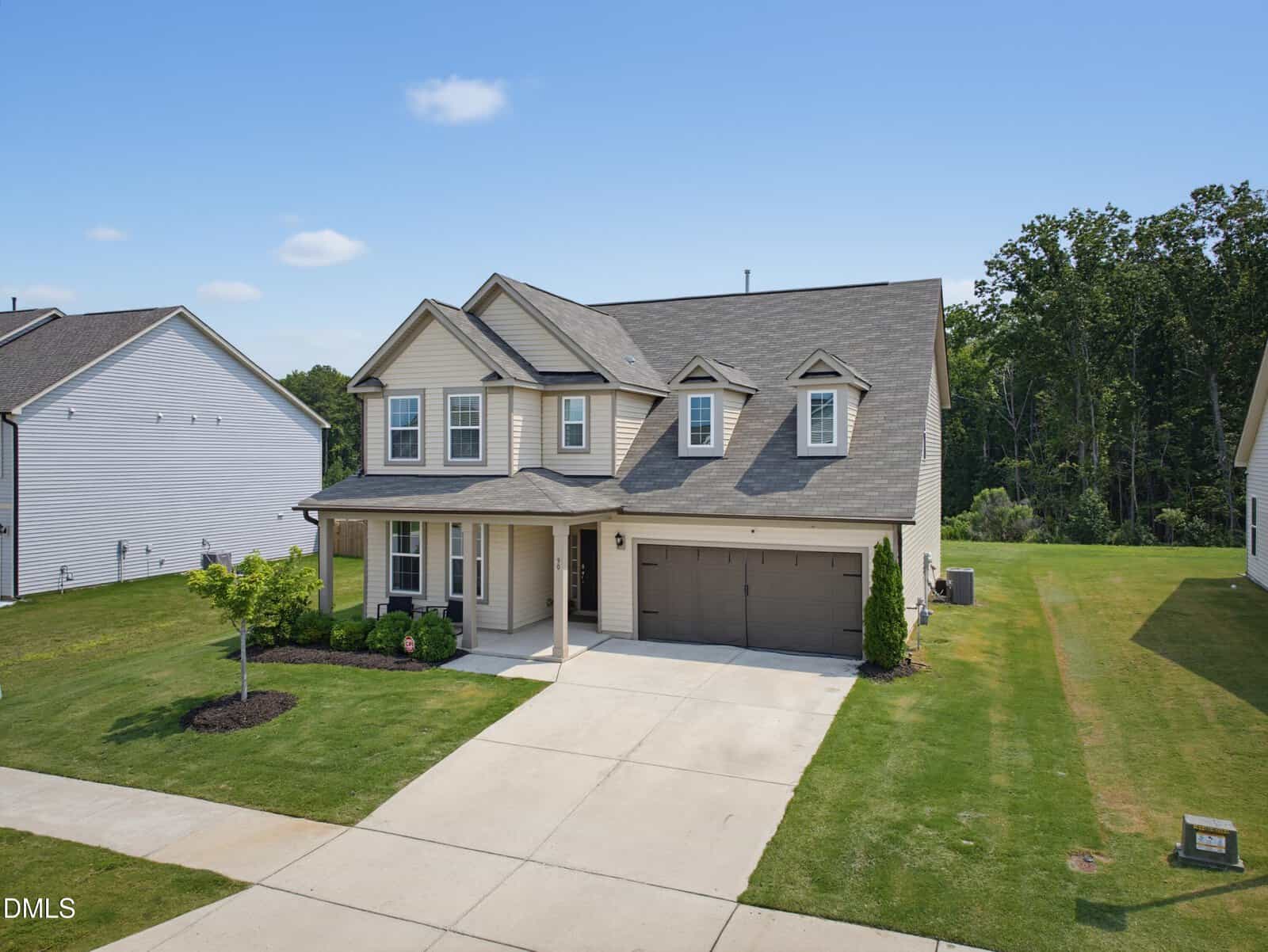 Two-story beige house with dark roof, attached three-car garage, covered front porch, and well-maintained lawn. The home is situated in a suburban neighborhood with similar houses and a wooded area in the background.