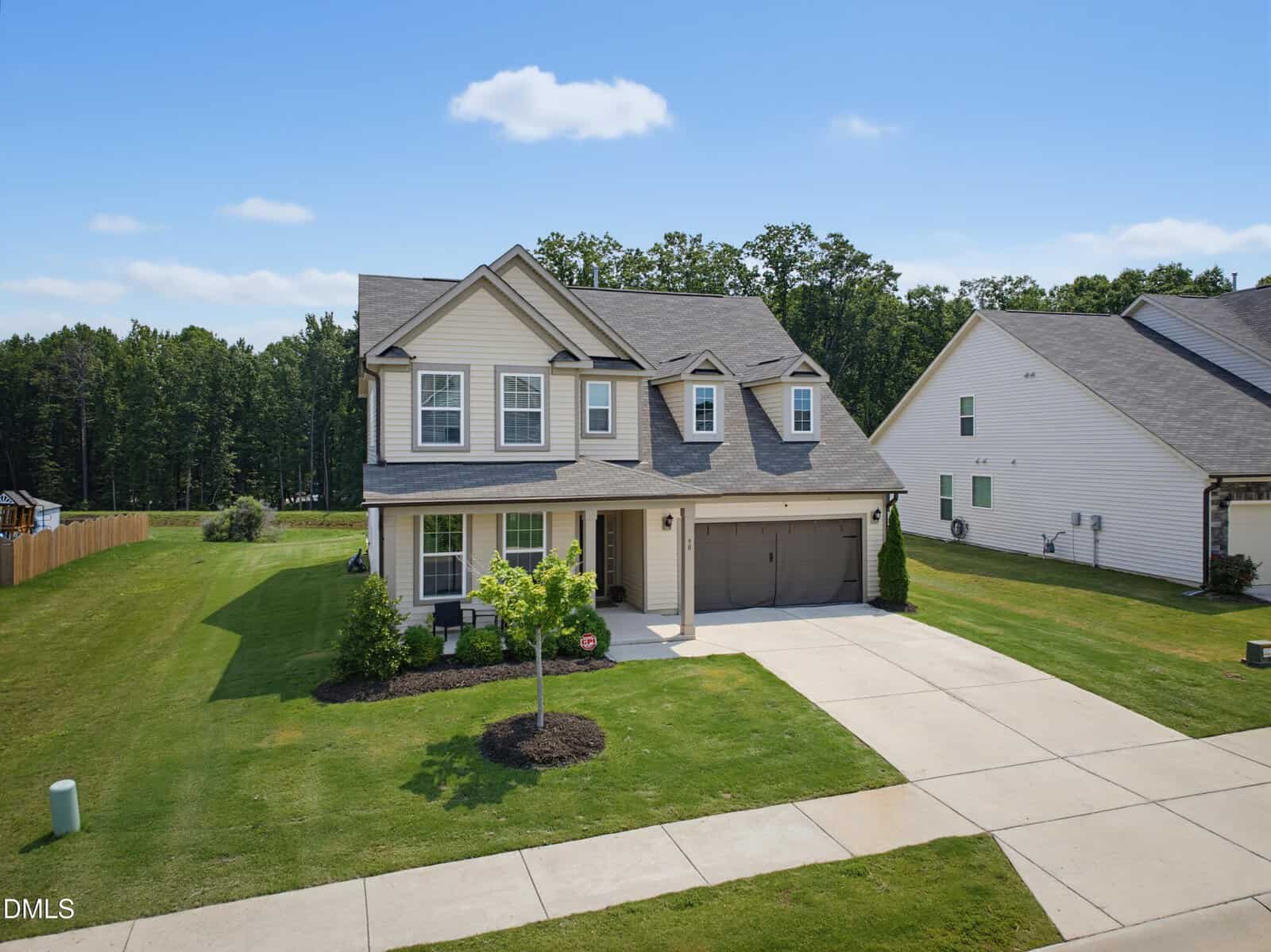 Two-story suburban house with gray siding, a covered front porch, and a three-car garage. The home is surrounded by a neatly mowed lawn, a small tree, and sits between similar houses under a clear blue sky.