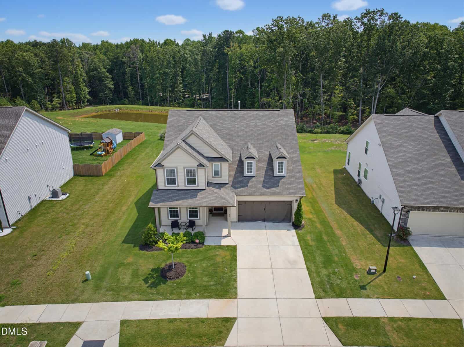 Two-story house with gray roof, front porch, and attached two-car garage. The home sits on a well-maintained lawn with a tree, surrounded by similar houses and backed by a wooded area under a clear sky.