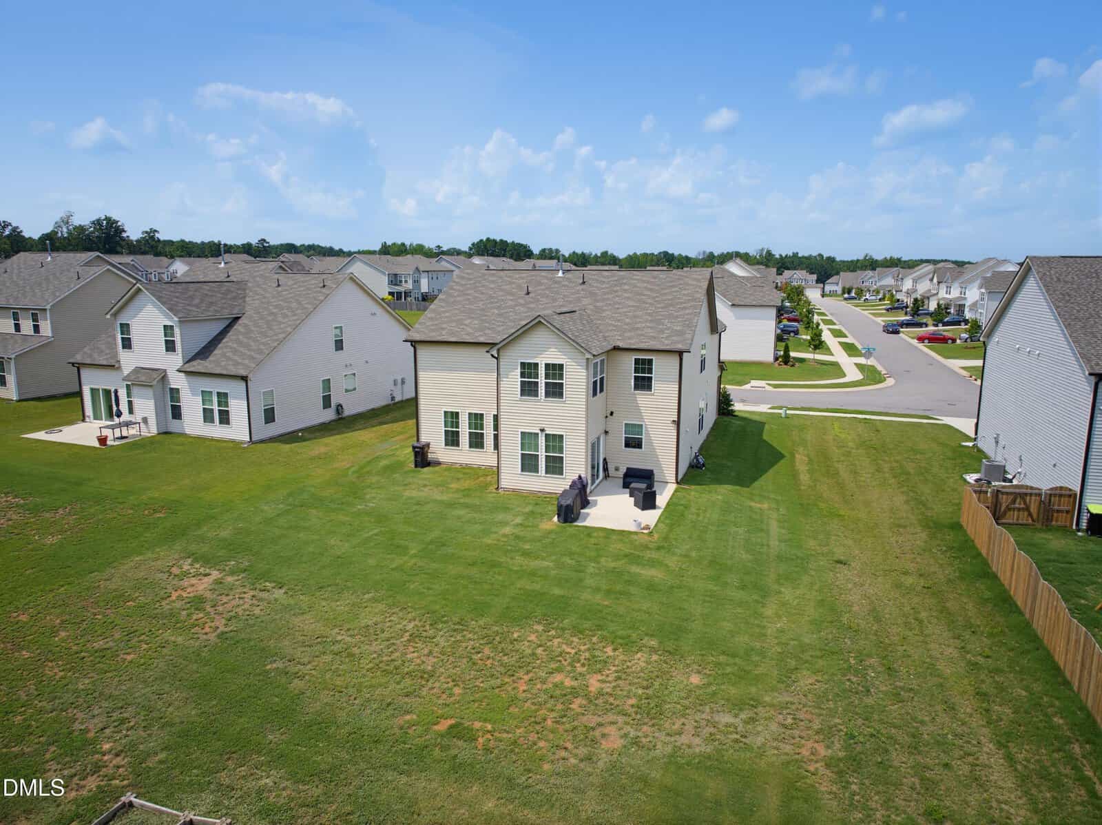 A two-story suburban house with a back patio is shown on a large grassy lawn, surrounded by similar houses and roads under a partly cloudy sky.