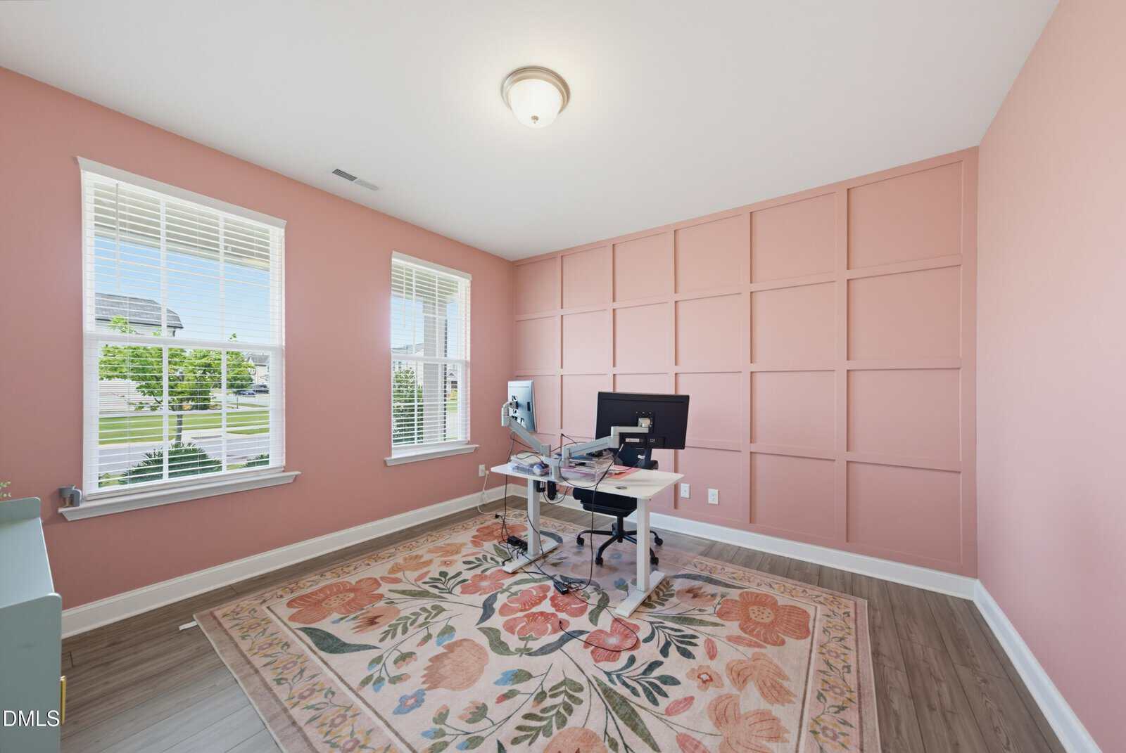 A home office with pink walls, a floral rug, and a white desk with dual monitors. Large windows let in natural light, and hardwood floors complete the bright, cozy space.