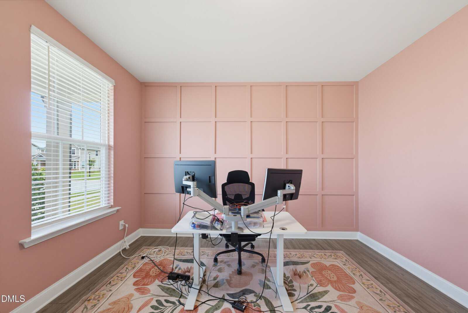 Modern home office with a pink paneled accent wall, two monitors on a white desk, office chair, floral rug, wood floor, and a large window letting in natural light.