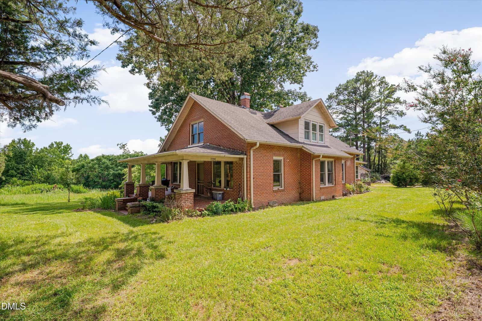 A two-story brick house with a covered front porch sits on a grassy lawn surrounded by trees and shrubs under a partly cloudy sky.