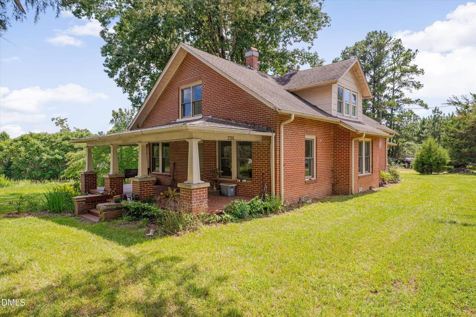 A two-story red brick house with a covered front porch sits on a grassy yard, surrounded by trees and shrubs under a blue sky with scattered clouds.
