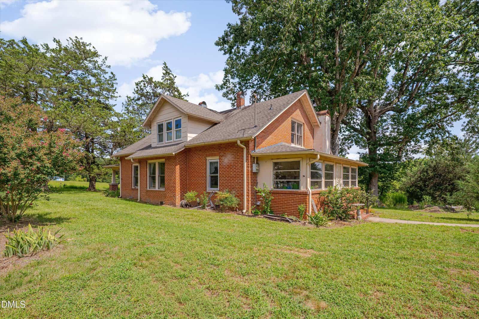Two-story brick house with a gabled roof and enclosed porch, surrounded by grass, trees, and landscaping on a sunny day with a partly cloudy sky.