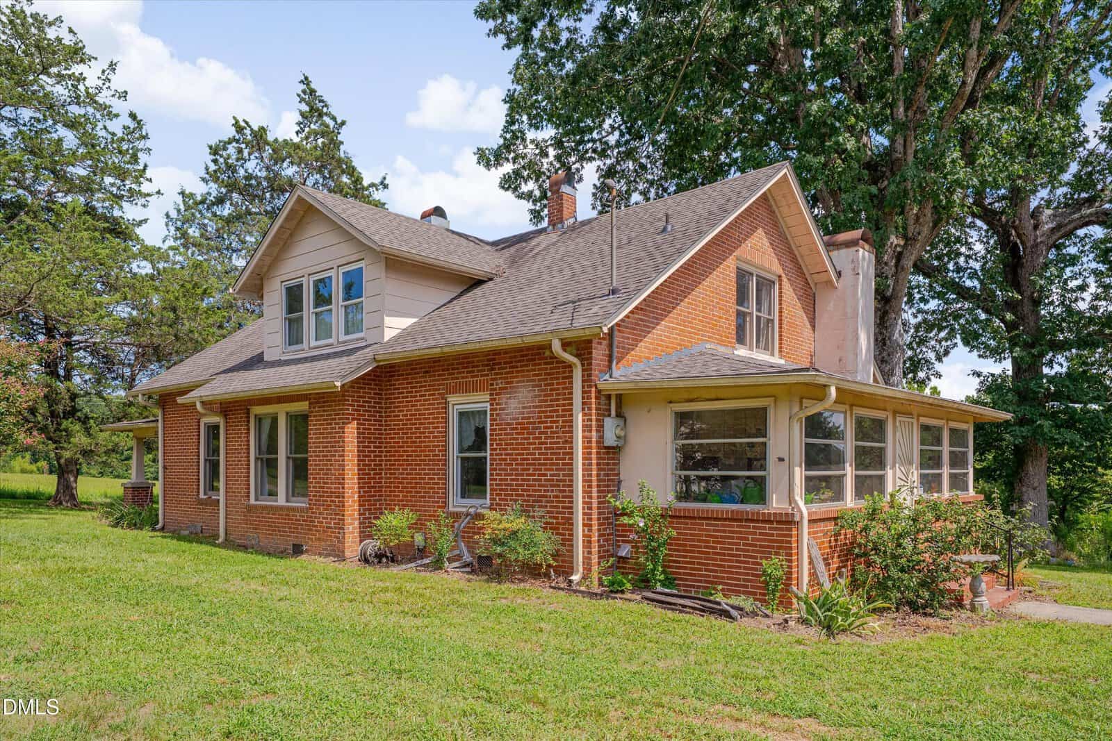 A brick house with a gabled roof, upper dormer windows, a sunroom, and a chimney, surrounded by green grass, shrubs, and tall trees on a sunny day.