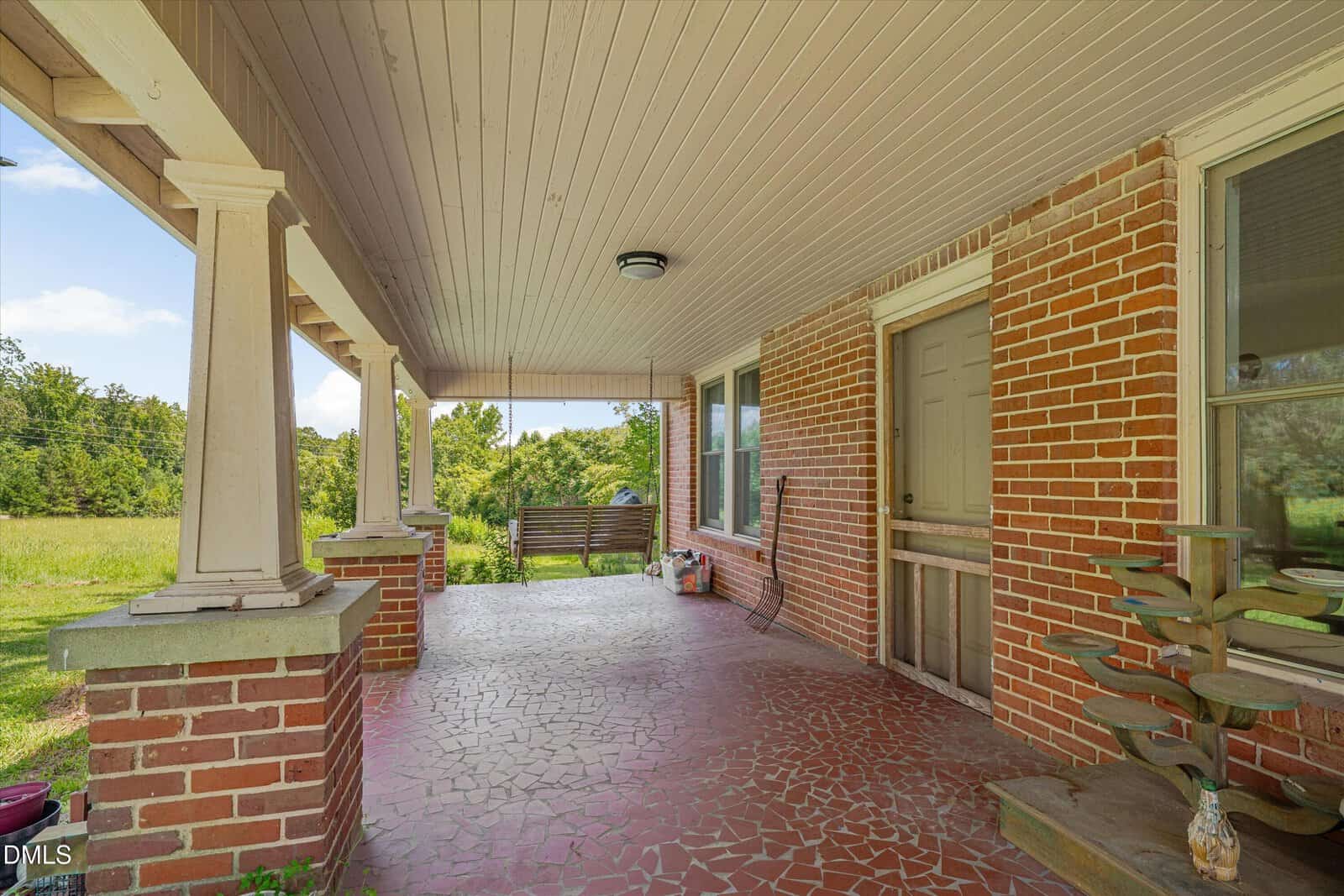 Covered brick porch with tiled floor, wooden swing, potted plants, and a wooden door. The porch overlooks a grassy yard and trees, with sunlight streaming in from the left.