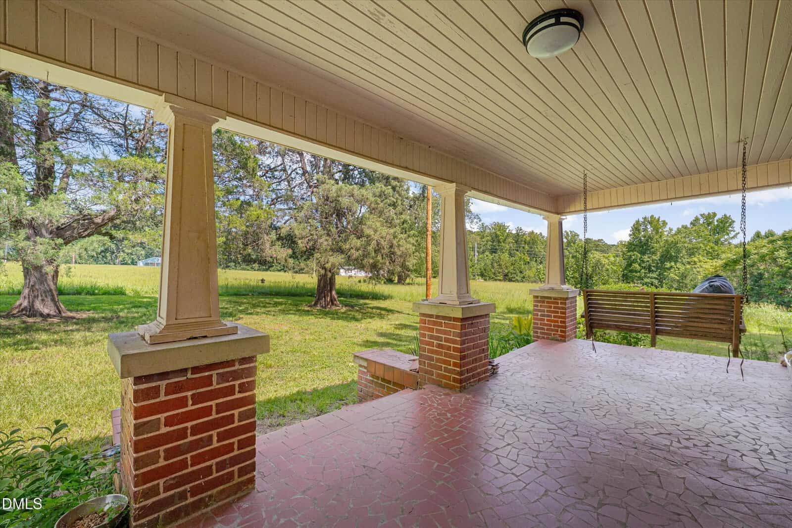 A covered porch with brick columns, a tiled floor, and a wooden swing overlooks a grassy yard with trees and a field in the background.