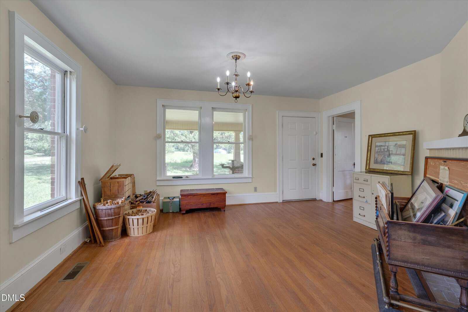 Bright room with hardwood floors, cream walls, large windows, a chandelier, baskets, a chest, a dresser with art, and a wooden desk with books and framed photos. Doorways lead outside and to another room.