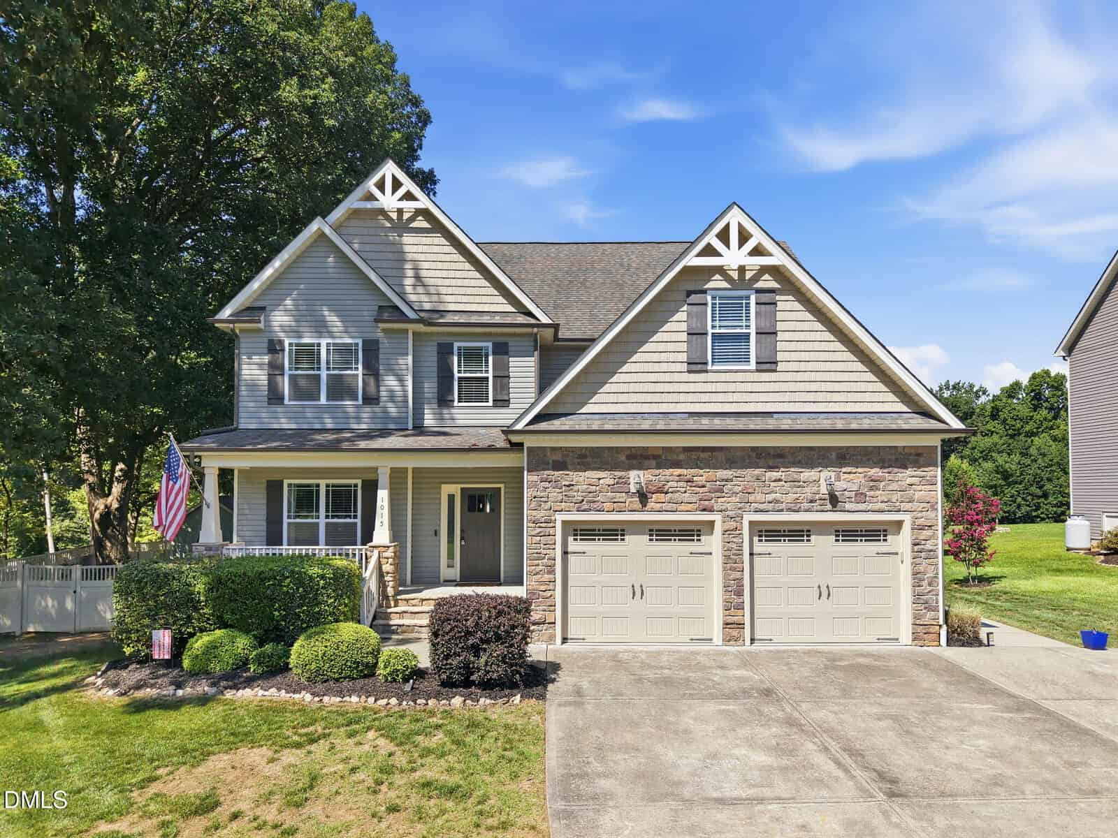 Two-story suburban house with beige siding, stone facade, and a double garage located at 1015 Bluebell Lane in Wake Forest. An American flag hangs on the front porch. The house is surrounded by a manicured lawn, bushes, and trees, with a clear blue sky above.