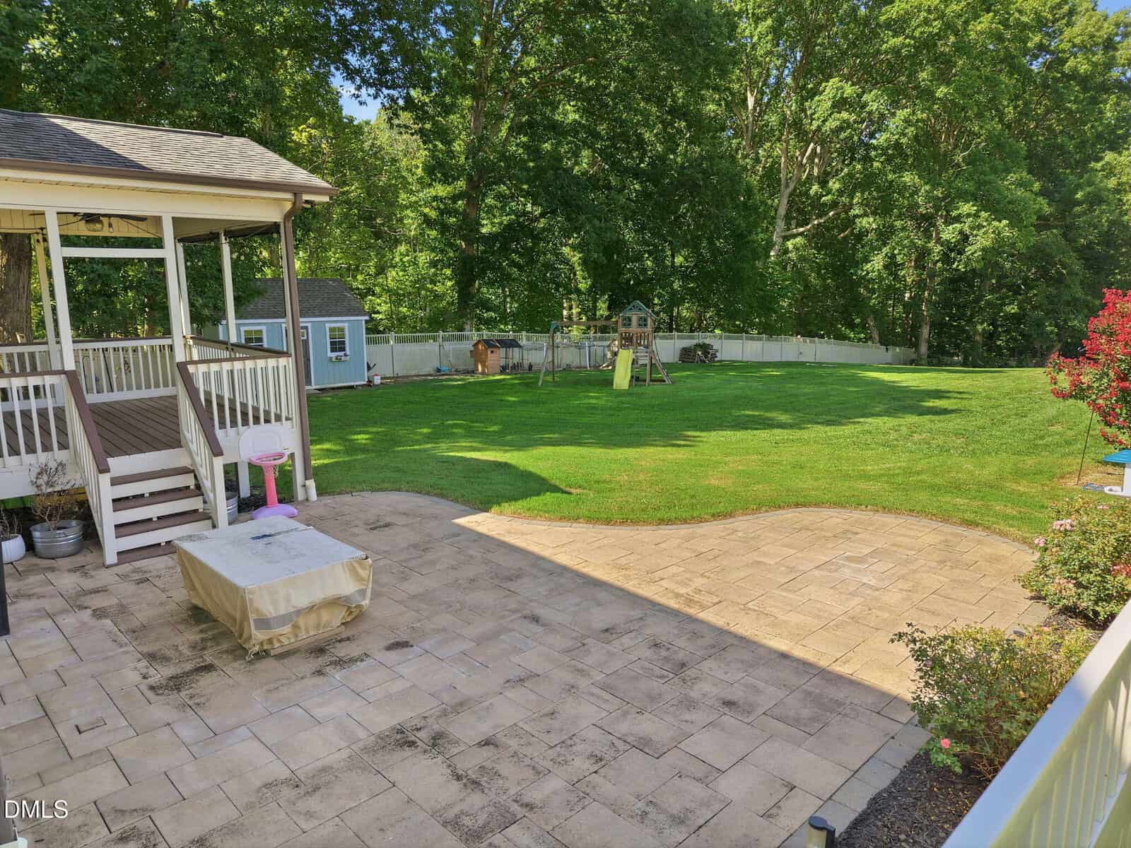 Spacious backyard with a gazebo, paved patio, lawn, white fence, shed, and children’s playset surrounded by trees under a clear sky.