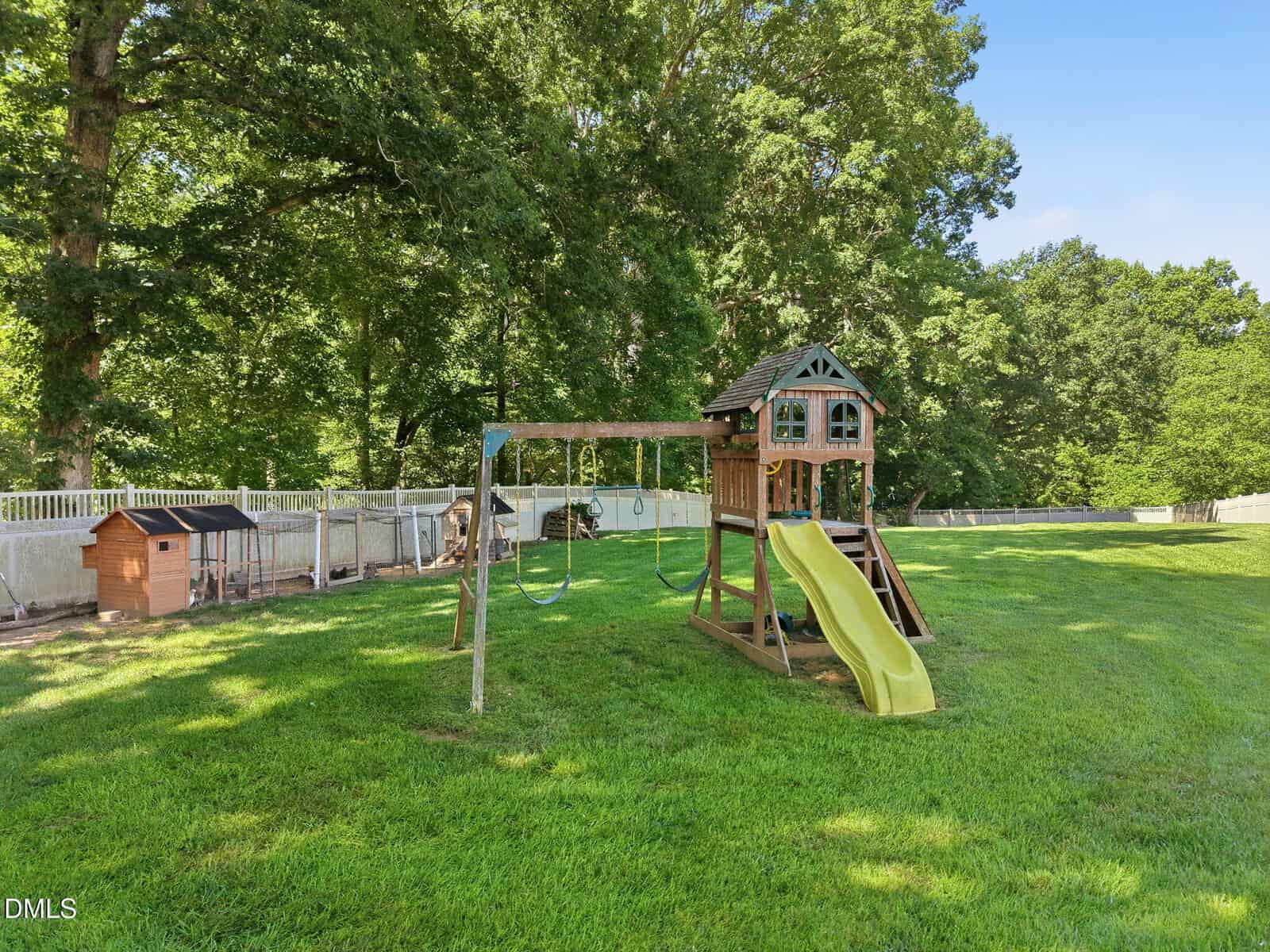 A wooden playset with swings, a yellow slide, and a small clubhouse stands on a grassy lawn. Several dog kennels line the white fence in the background, and tall trees provide shade. The sky is clear and blue.